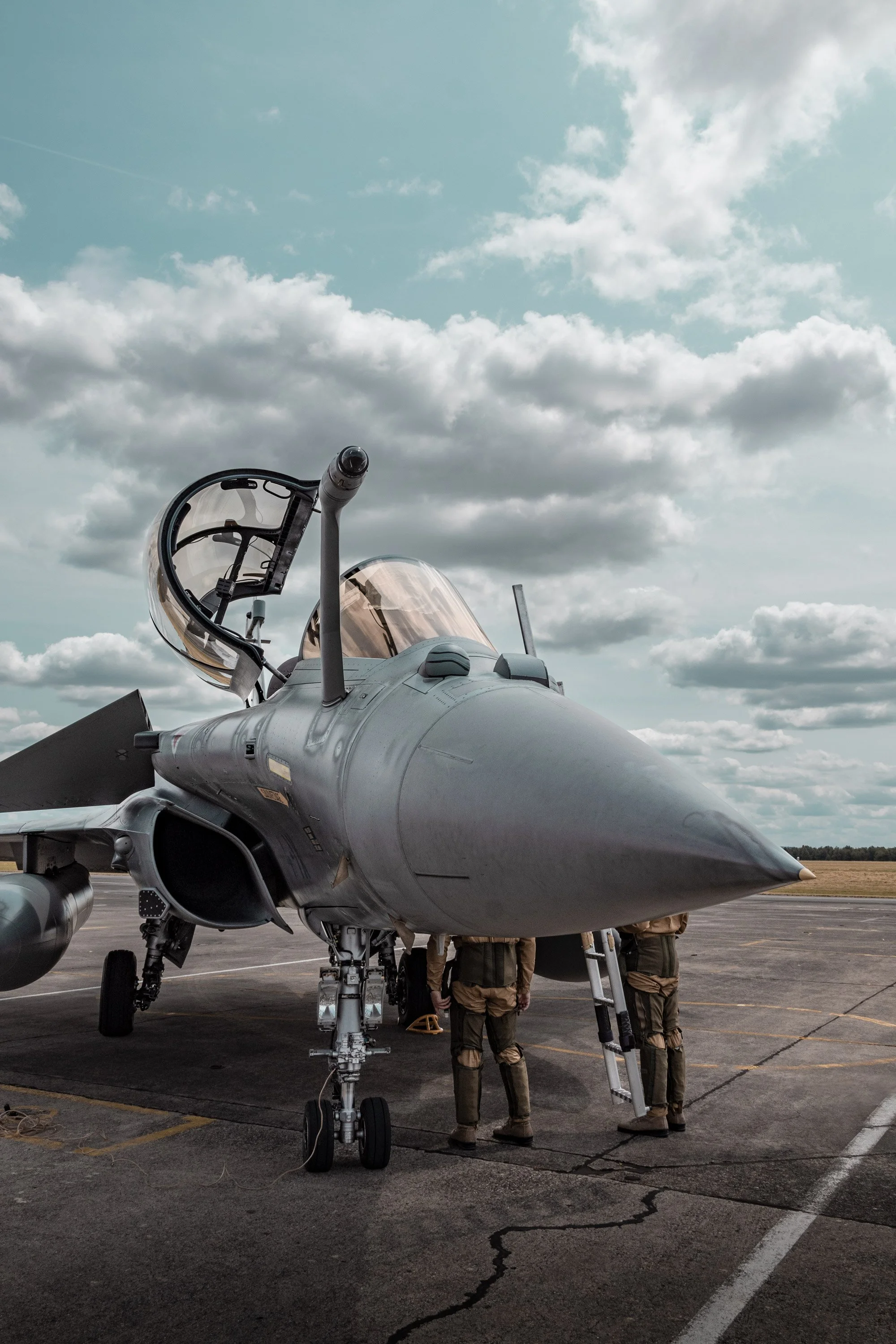 Un avion de chasse Rafale Français au sol, avec des militaires en formation autour, sous un ciel nuageux.