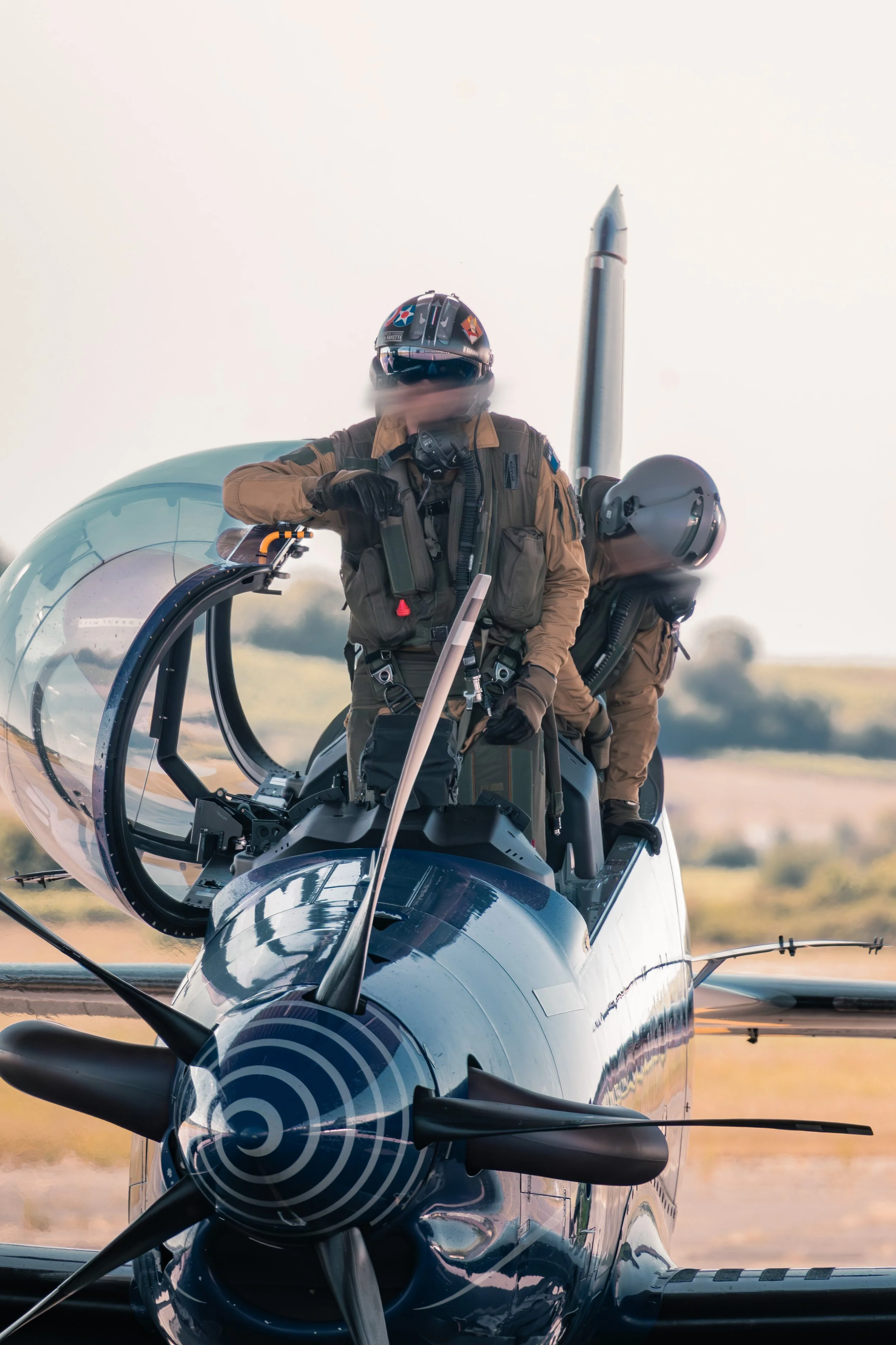 Deux pilotes dans un avion de chasse PC21 Français, prêt à décoller, portant des casques et des combinaisons de vol de couleur kaki, avec un environnement aéroport en arrière-plan. Base aérienne 709 de Cognac