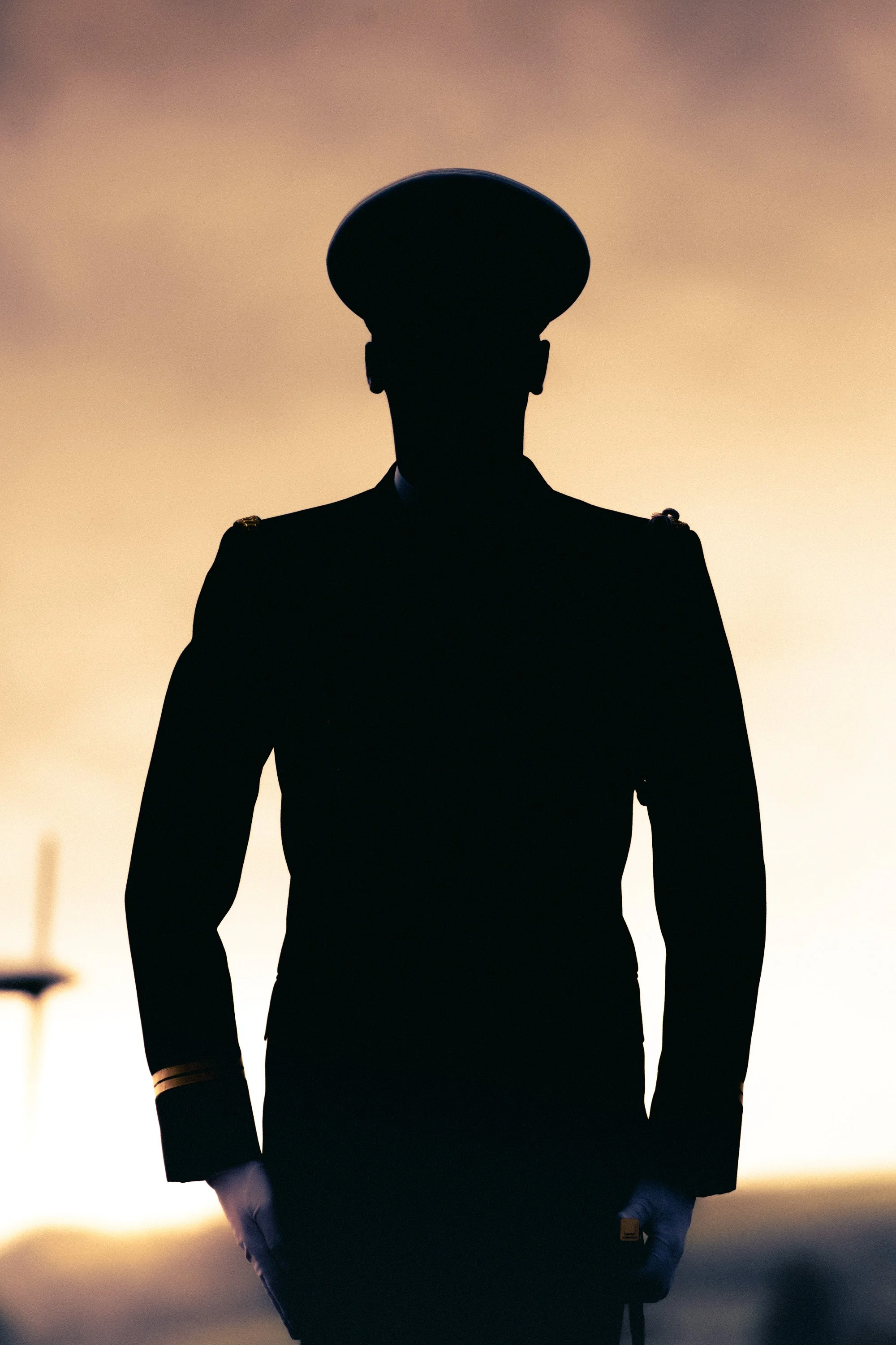 Silhouette d'une personne en uniforme militaire, portant un chapeau, contre un ciel en dégradé de couleurs au coucher du soleil.
