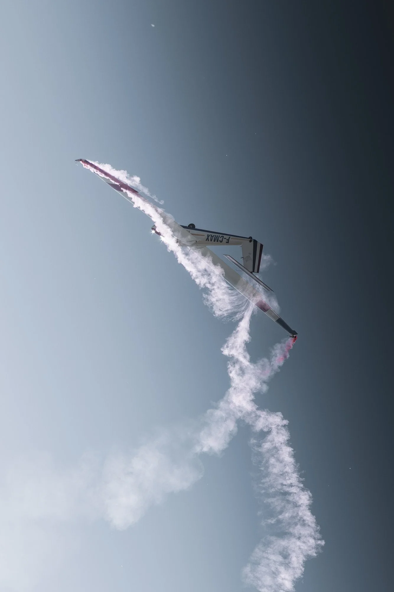Un avion planeur de voltige aérienne effectuant une manœuvre en vol, laissant derrière lui une traînée de fumée blanche et rose contre un ciel clair.