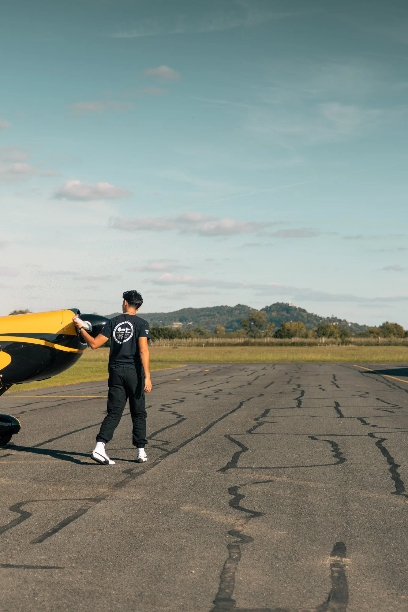 Un pilote de voltige se prépare à monter dans un avion léger de couleur jaune CAP 10 et noire dans un aérodrome Villeneuve sur Lot, avec un paysage rural en arrière-plan et un ciel partiellement nuageux.
