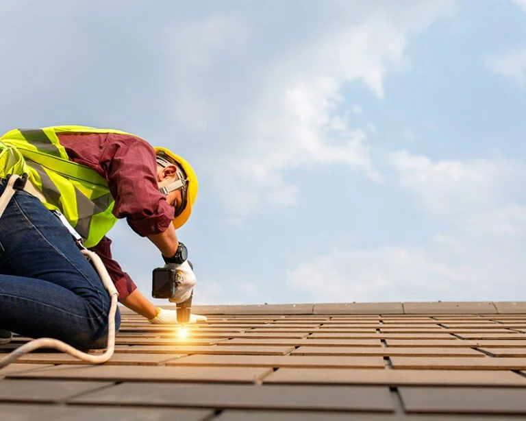 Een bouwvakker in een gele veiligheidshelm en hoge zichtbaarheidsvest die met een boormachine een houten dakplaat bevestigt, onder een partly bewolkte blauwe lucht.