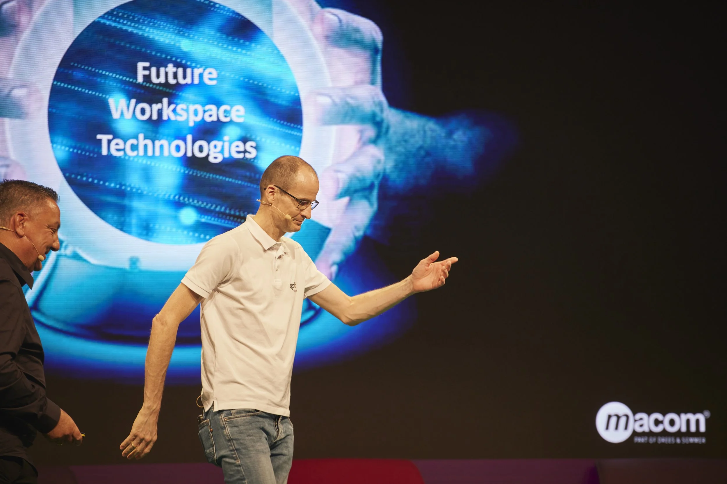 Zwei Männer auf einer Bühne bei einer Präsentation, im Hintergrund ein großer Bildschirm mit der Aufschrift 'Future Workspace Technologies'.