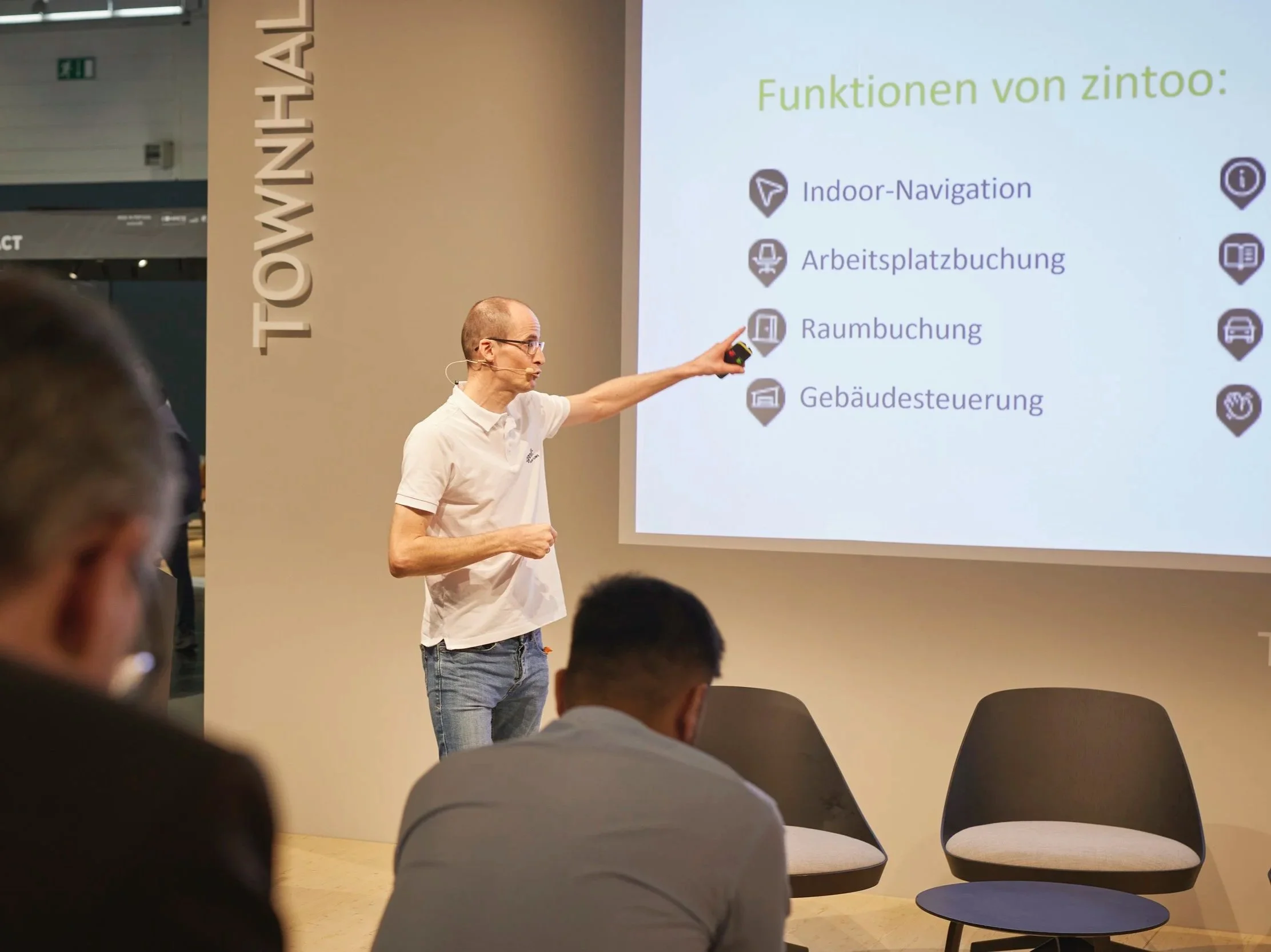 Ein Mann präsentiert auf einer Bühne eine Folie mit Text und Symbolen. Er zeigt auf die Folie, die die Funktionen von zintoo beschreibt, darunter Indoor-Navigation, Arbeitsplatzbuchung, Raumbuchung und Gebäudesteuerung.