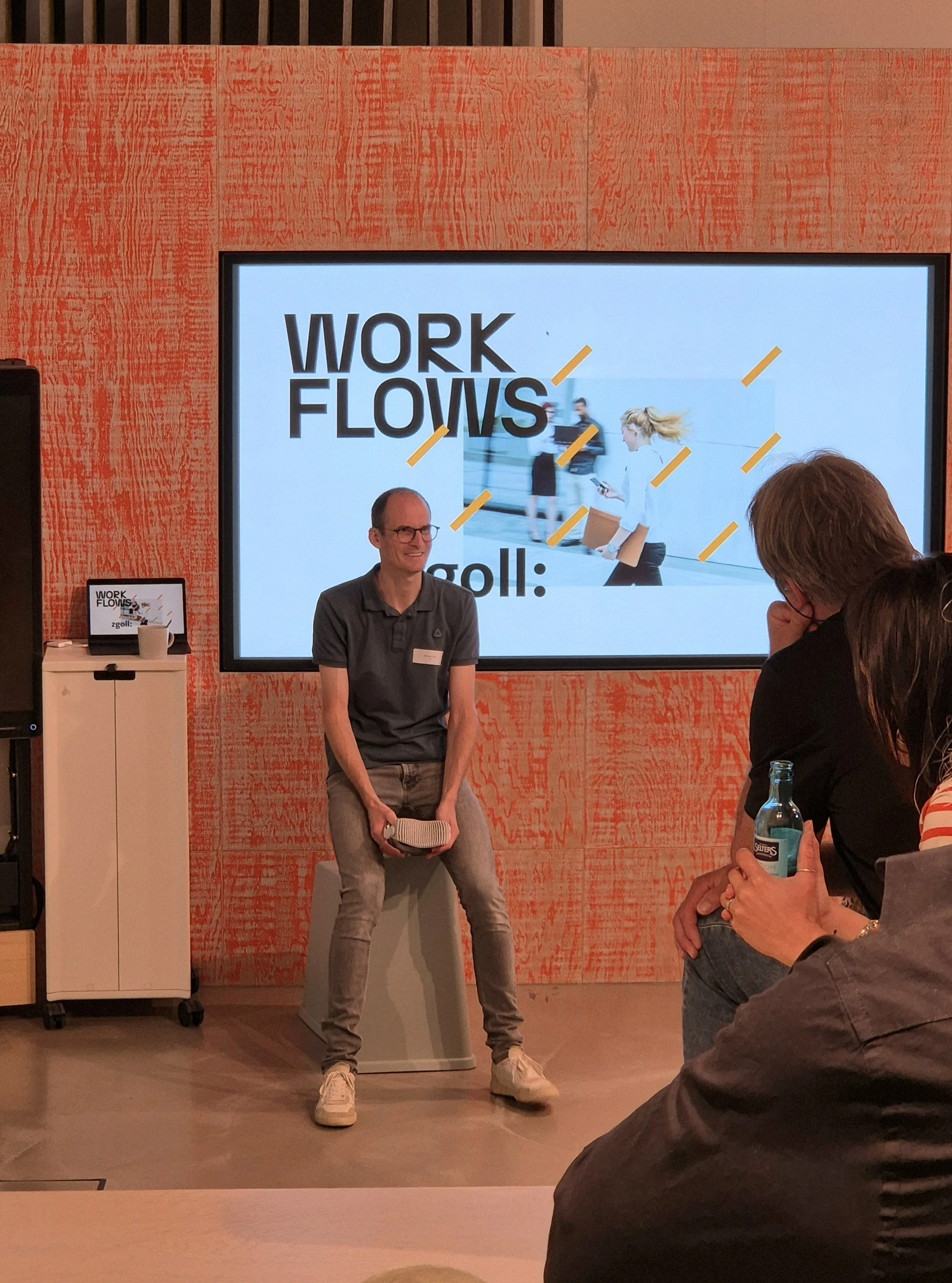 Ein Mann sitzt auf einem Hocker vor einem großen Bildschirm bei einer Präsentation mit dem Thema "Work Flows", während er spricht. Im Vordergrund sitzen einige Zuhörer, einer hält eine Flasche. Die Präsentation läuft an einer orange gestalteten Wand.