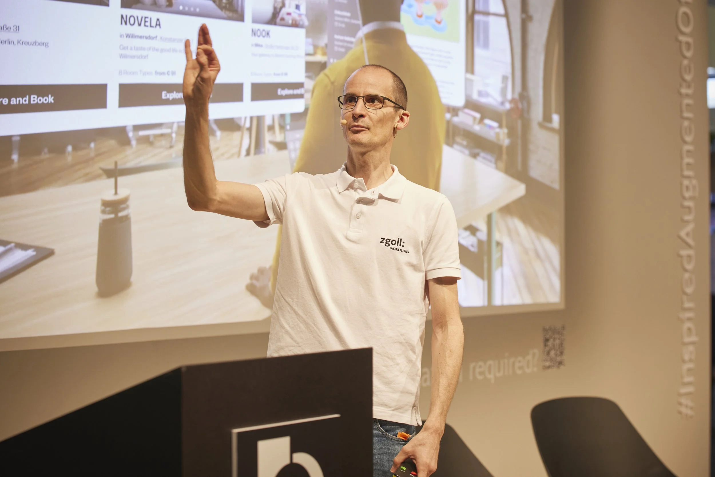 Man während einer Präsentation, der auf eine digitale Tafel zeigt, an einem Vortrag, mit einem Mikrofon am Ohr, trägt ein weißes Polo-Shirt mit dem Logo 'zgoll'.