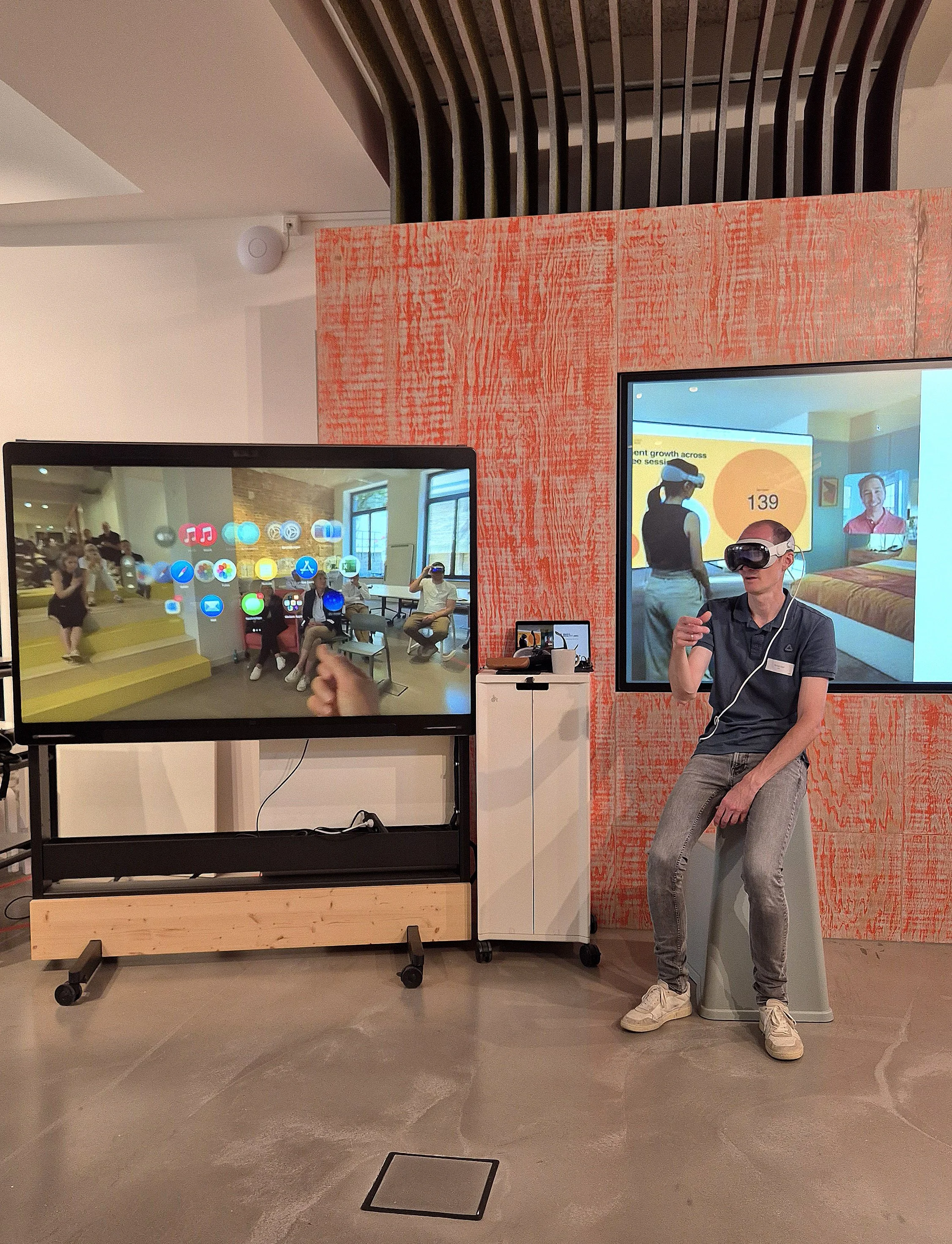 Eine Person sitzt mit einer VR-Brille auf einem Stuhl vor zwei großen Bildschirmen, die Virtual-Reality-Szenen zeigen, im Hintergrund eine orange Wand mit Mustern.