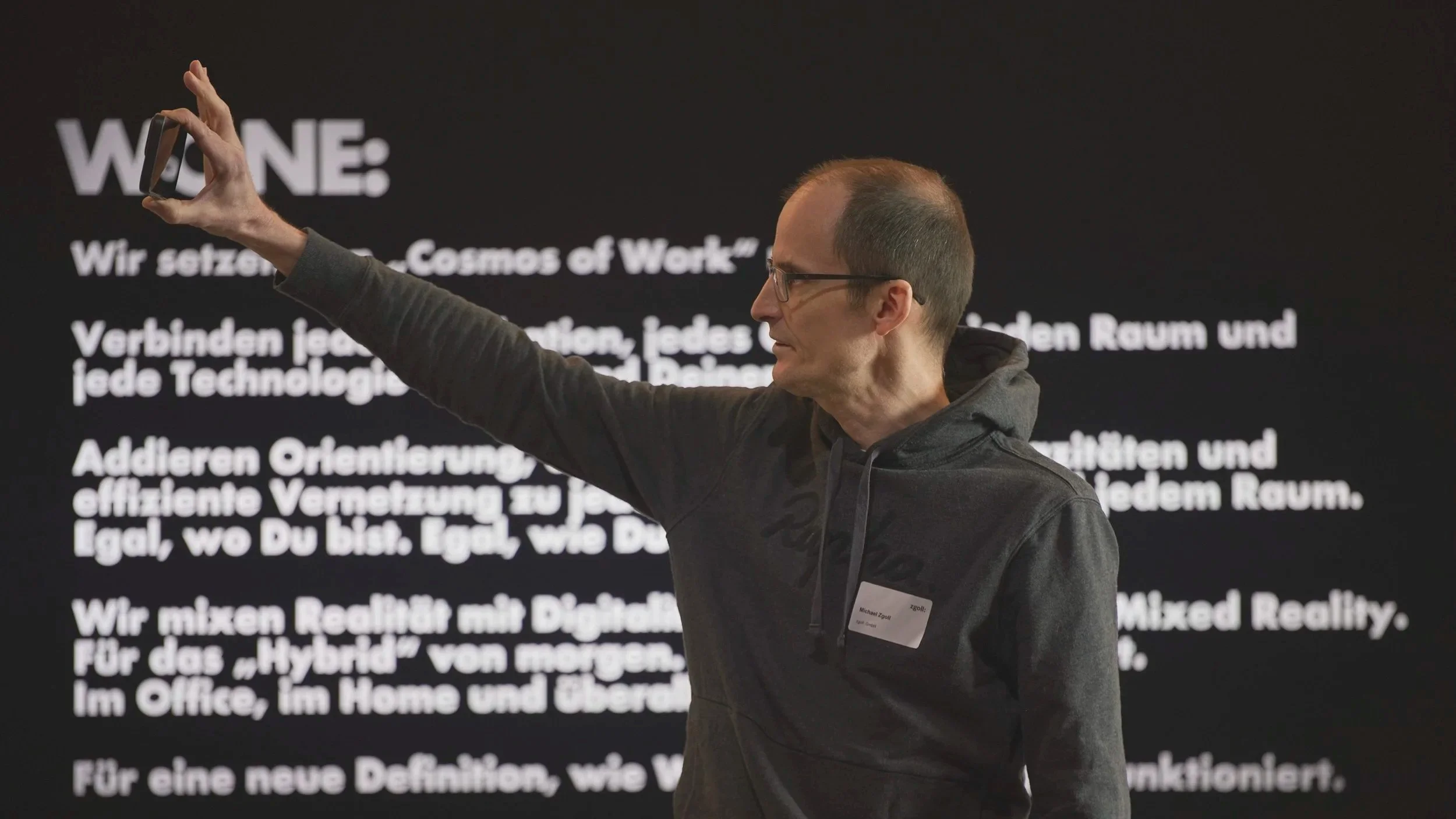 Mann in Brille und Hoodie macht ein Selfie vor einem schwarzen Präsentationsschirm mit deutschem Text.