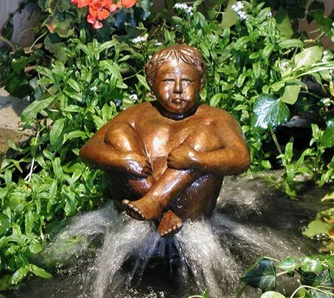Bronze statue of a young child sitting with legs crossed in a fountain, surrounded by green plants with some red flowers in the background.