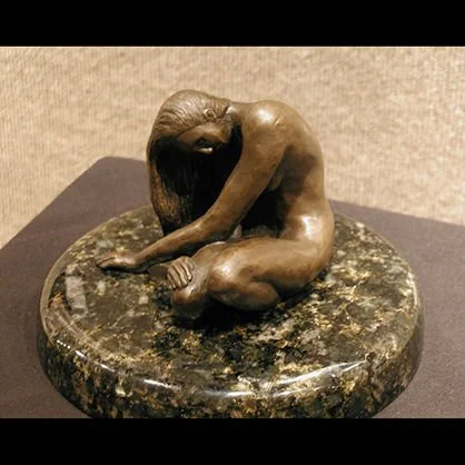 Bronze sculpture of a girl sitting with her head resting on her knees, on a round marble base.