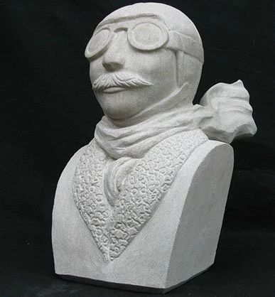 Stone bust of a man with a mustache, wearing goggles and a scarf, with a textured vest.