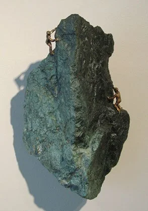 Two climbers scaling a large, jagged rock formation recorded indoors with a plain wall background.