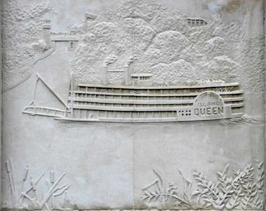 A white wall relief sculpture of a cruise ship named 'Island Queen' sailing on water with plants in the foreground.