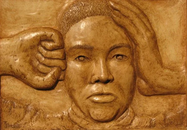 A carved wood sculpture of a person with short hair, making a fist with one hand near their head and the other hand resting on their head.
