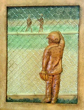 Child in baseball uniform standing with glove, looking at players on the field behind a chain-link fence.
