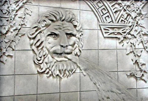 A stone carvings of a lion's face with water flowing from its mouth, surrounded by decorative elements on a wall.
