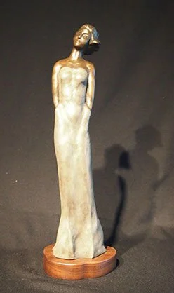 Sculpture of a stylized, elongated figure of a woman with short hair, standing on a wooden base, against a dark background.