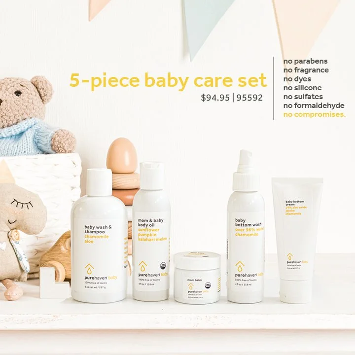 A five-piece baby care set displayed on a white surface with a plush teddy bear, a cloth doll, and baby accessories in the background. The set includes baby wash and shampoo, mom and baby body oil, baby bottom wash, mom balm, and baby cream, all labeled with natural ingredients like chamomile, aloe, sunflower, and calahari melon.