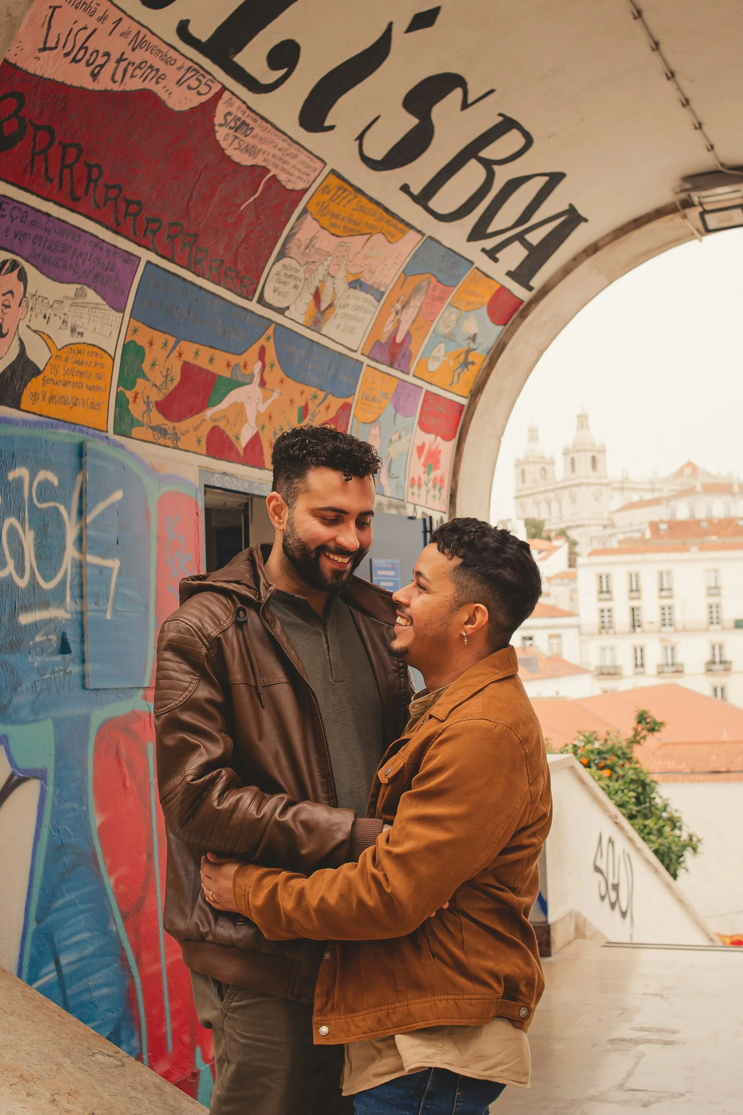 Two masculine-presenting individuals of color embrace and smile beneath an artsy archway.