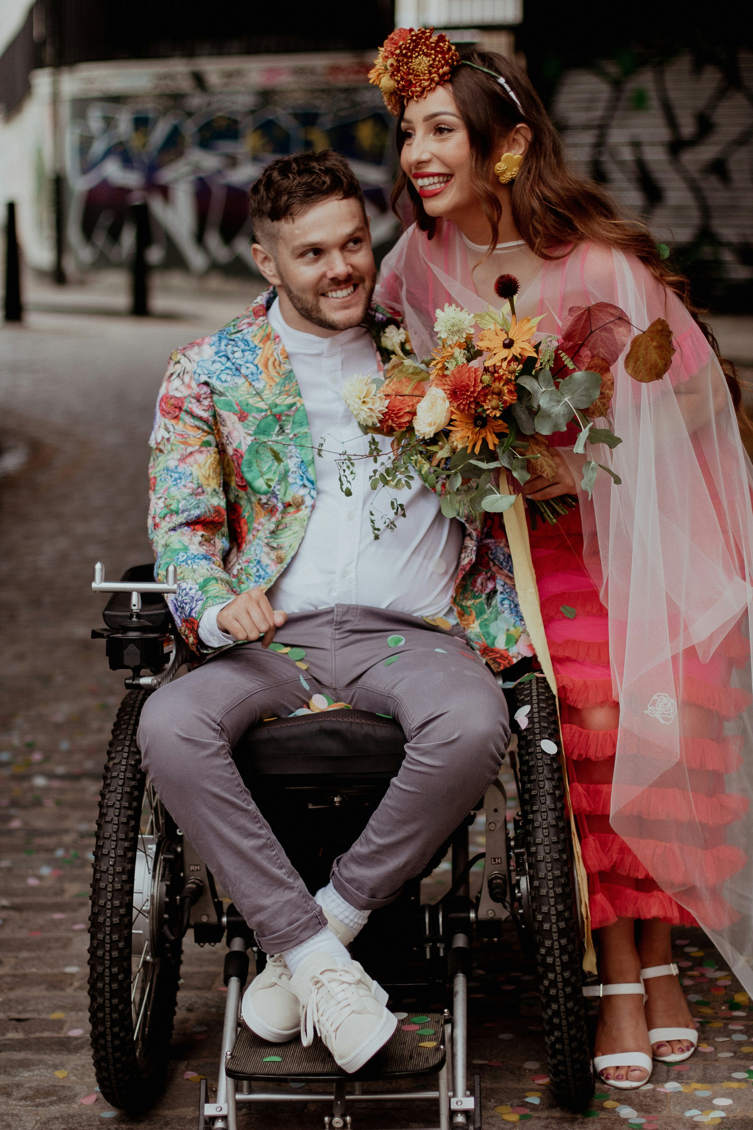 A masculine-presenting individual in a wheelchair and their feminine-presenting partner smile and hold each other in celebration.