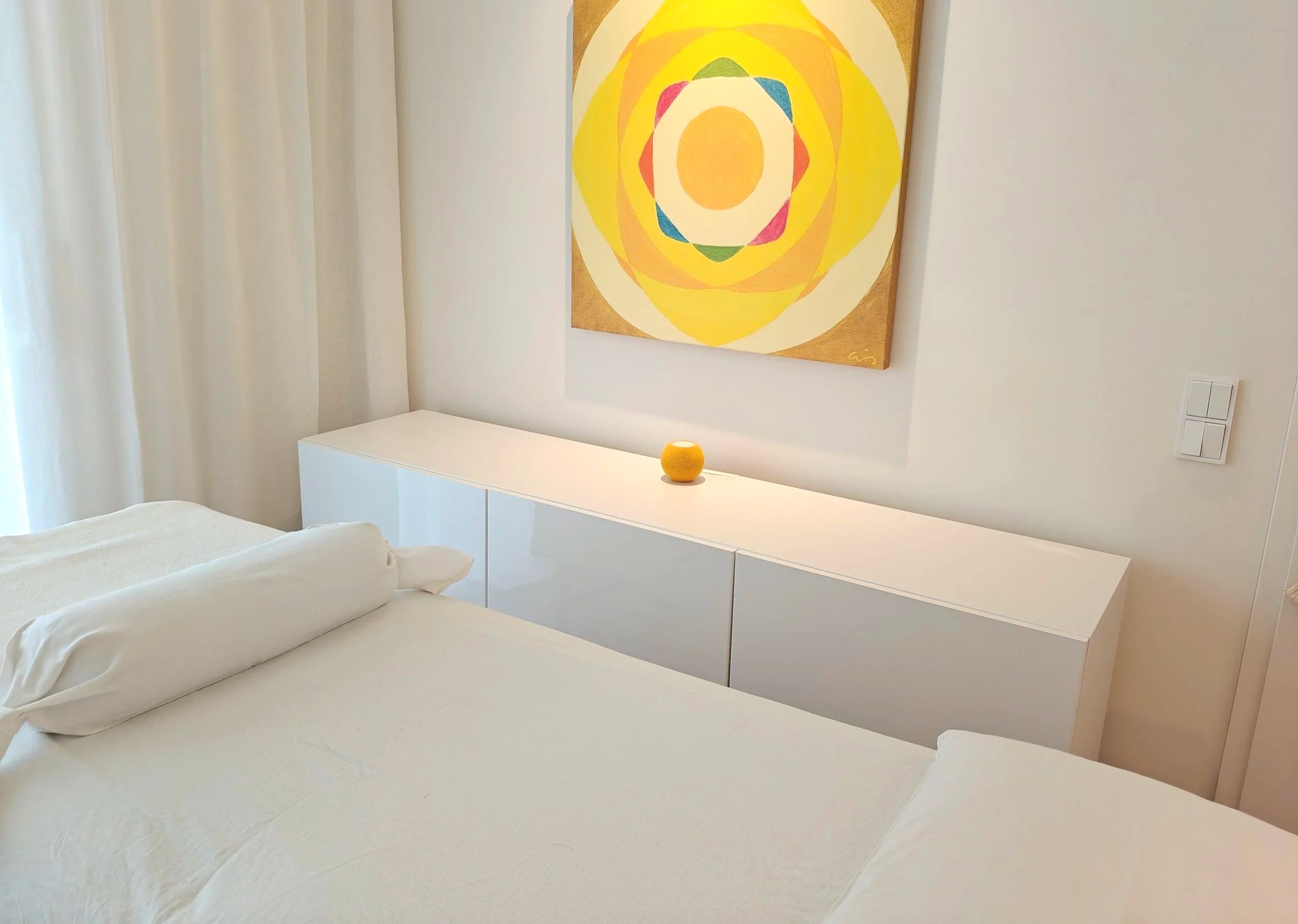 Ein Praxiszimmer mit Behandlungsliege, einem weißen Sideboard, einem gelben Lautsprecher und einem bunten, geometrischen Wandbild.