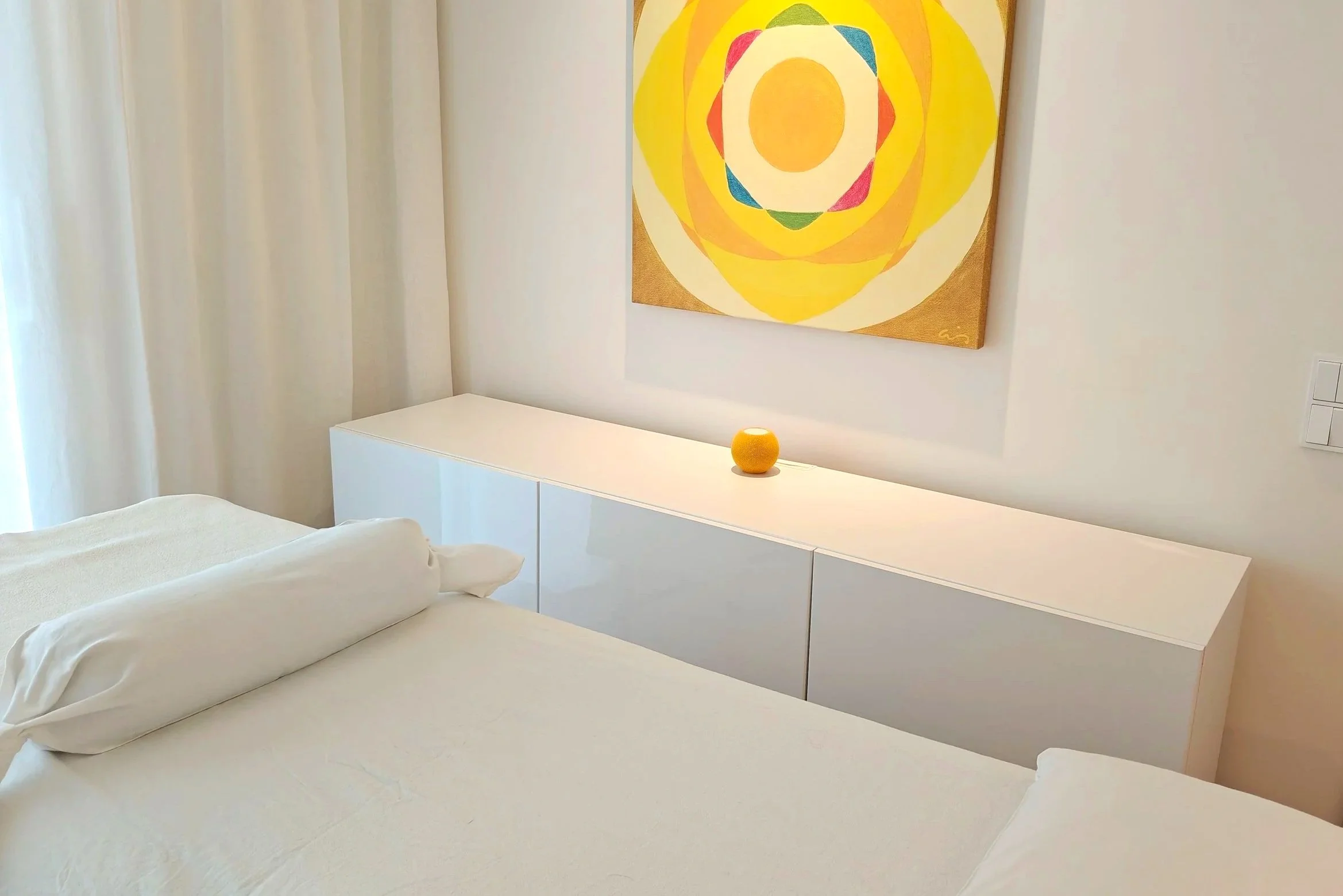 Ein Praxiszimmer mit Behandlungsliege, einem weißen Sideboard, einem gelben Lautsprecher und einem bunten, geometrischen Wandbild.