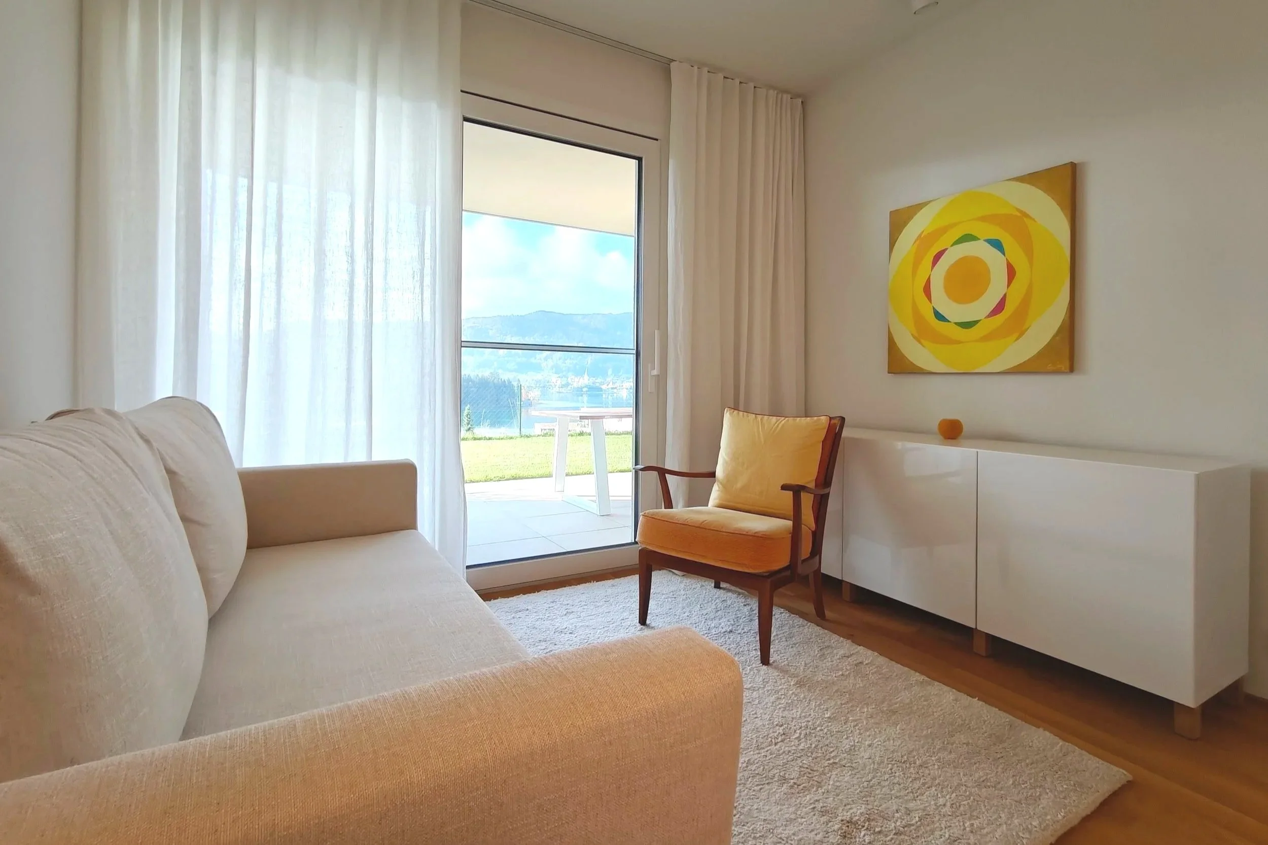 Gemütliches Praxiszimmer mit großem Fenster, Blick auf den Terrasse, die Natur und See, beige Couch, orange-armiger Sessel, weiße Kommode, bunte geometrische Wandkunst.