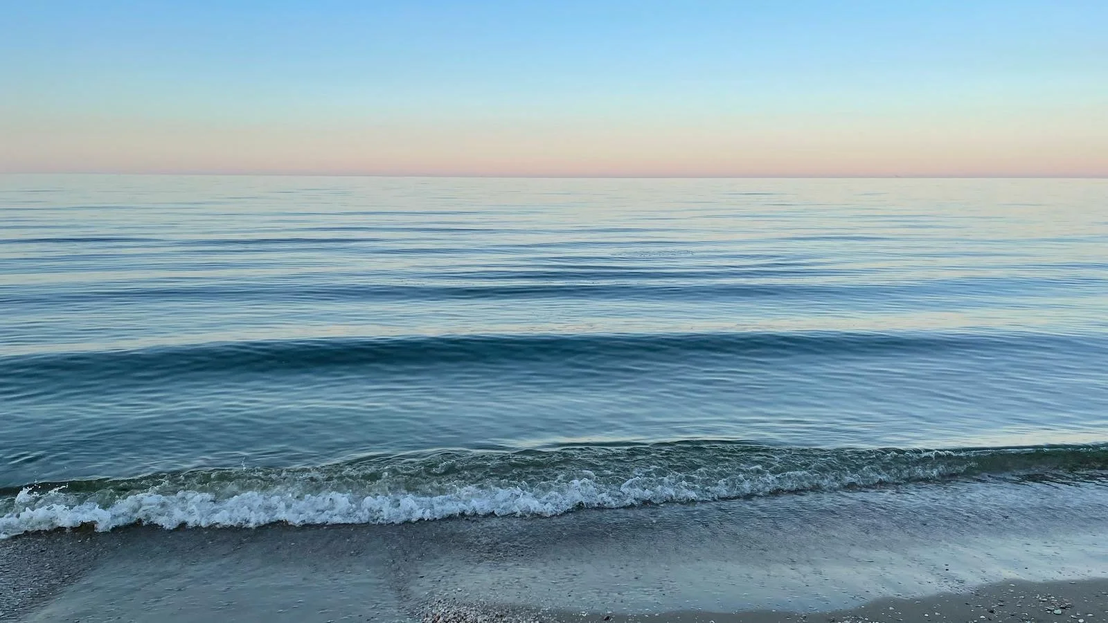 Das Bild zeigt eine ruhige Meeres- oder Ozeanszene mit sanften Wellen am Strand unter einem Morgendämmerungshimmel in Pastellfarben.