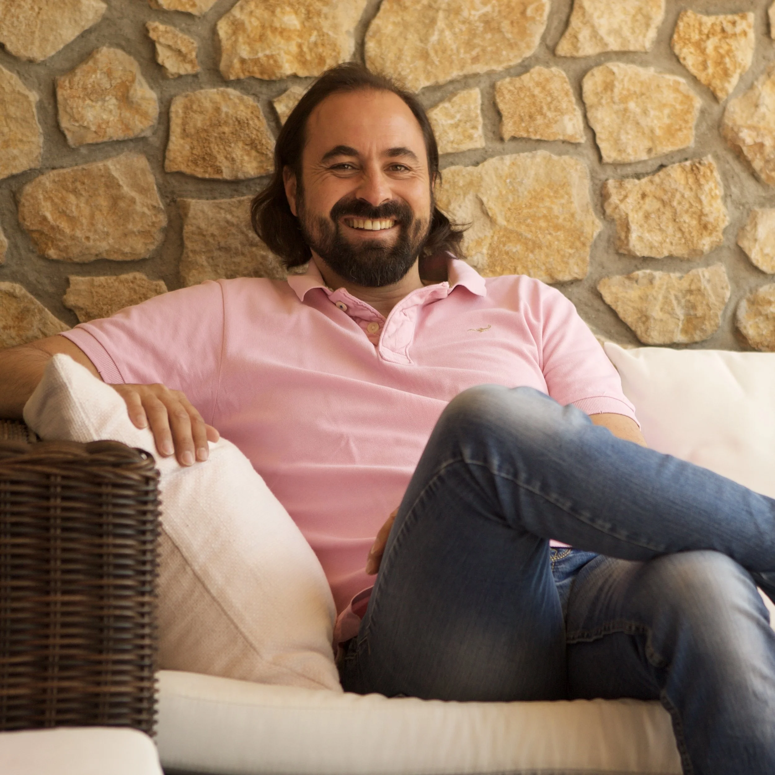 Ein lachender Mann mit dunklem Haar und Bart, trägt ein pinkes Polo-Shirt, sitzt auf einem weißen Couch vor einer Steinmauer.