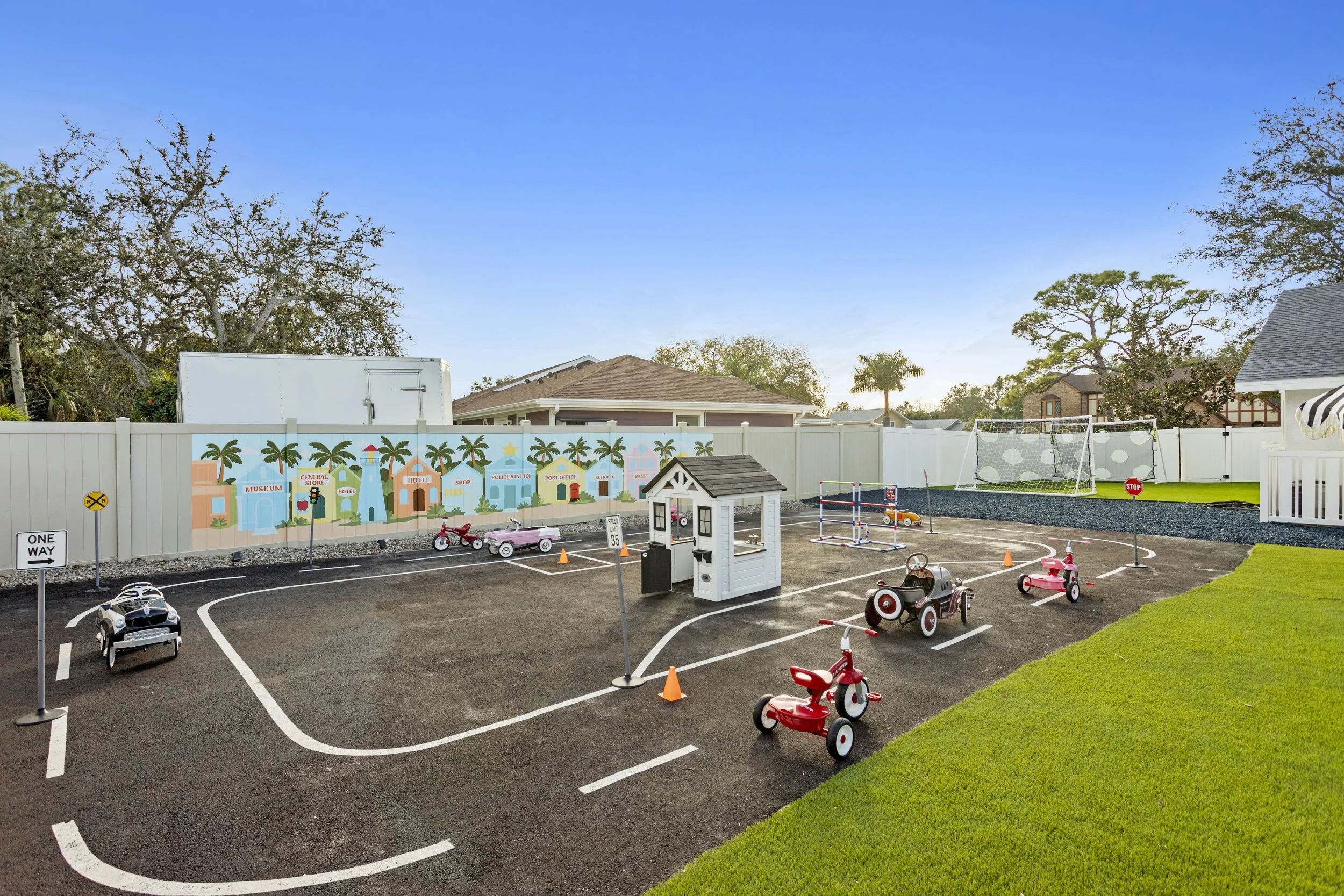 Children's outdoor playground with toy cars, small traffic signs, a miniature house, and a painted mural of a town with buildings, palm trees, and a lighthouse, enclosed by a white fence, with a grassy area and trees in the background.