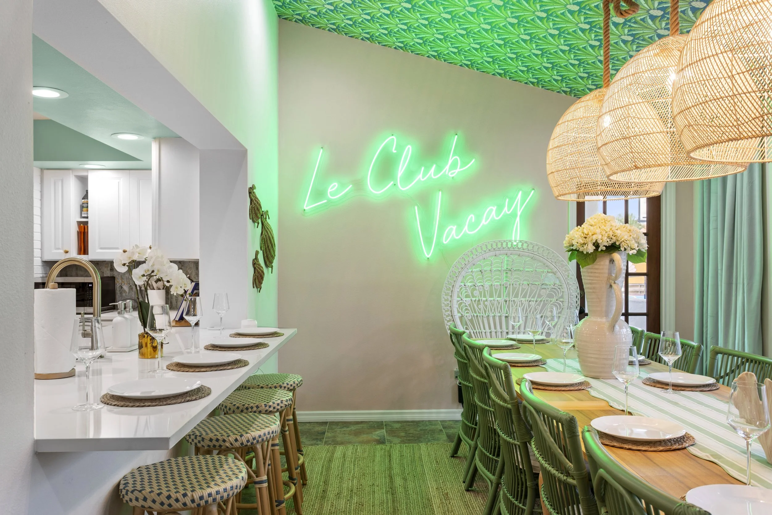 A dining area with a neon sign reading 'Le Club Vacay' on the wall, a large white vase with white flowers on the table, wicker pendant lights, and a long wooden table set with plates, glasses, and woven placemats; light green accents and curtains in the background.