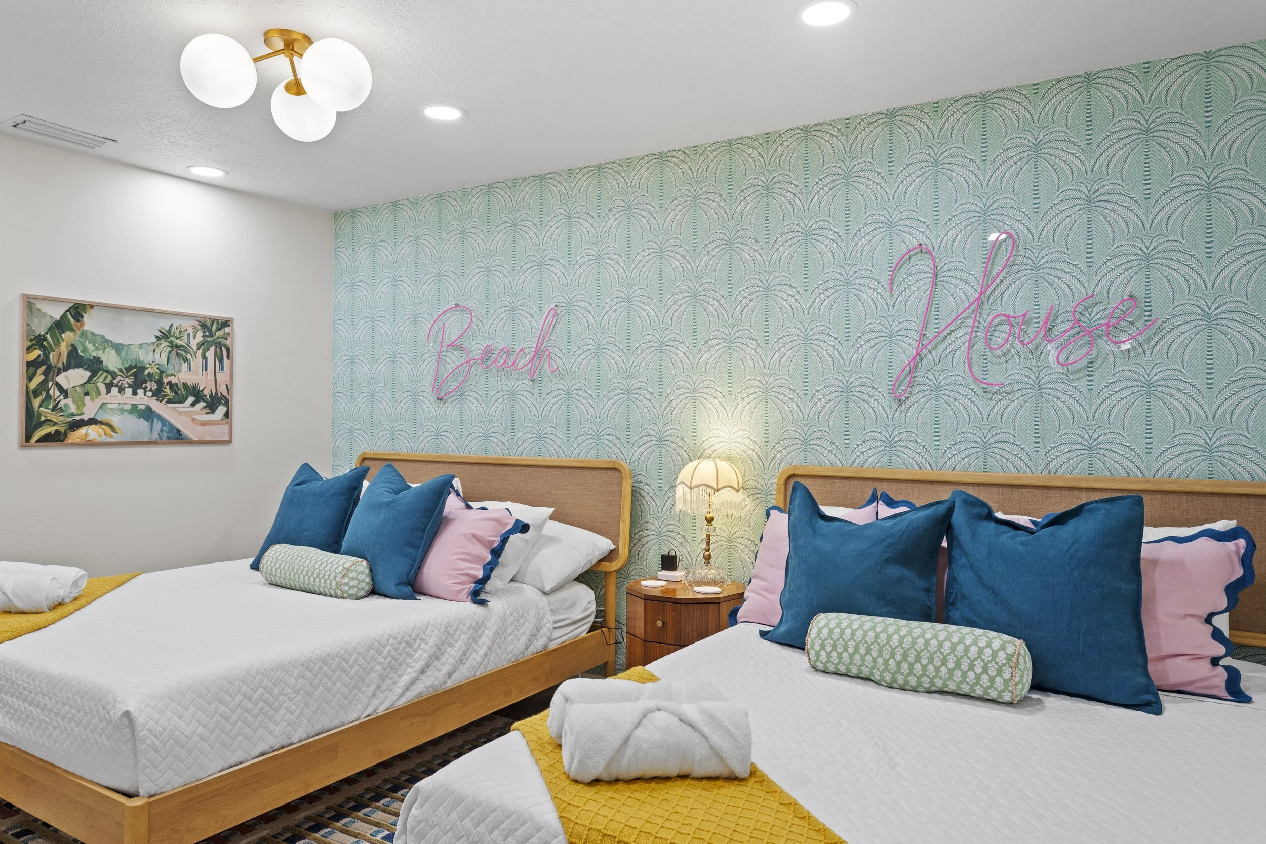 Two beds in a beach-themed hotel room with white bedding, blue and pink pillows, a small bedside table with a lamp, a towel, and a painted tropical landscape on the wall. Behind the beds, a blue patterned wallpaper features a neon pink sign reading "Beach House."
