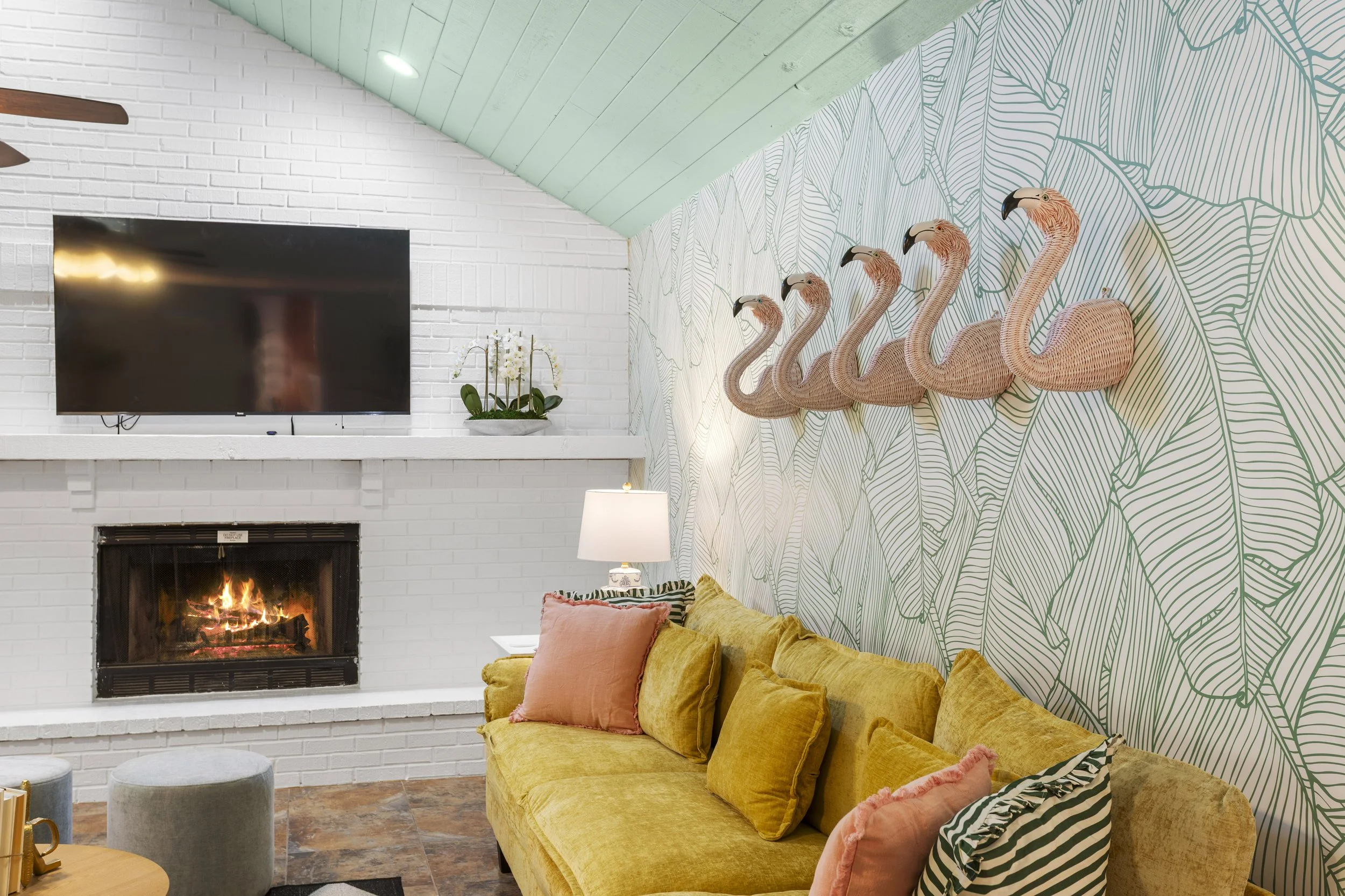 Living room with yellow velvet sofa, pink and striped throw pillows, a white wall-mounted fireplace with a fire, a mounted TV, a lamp, a white shelf with an orchid plant, and wall decor of pink flamingos on a leafy wallpaper background.