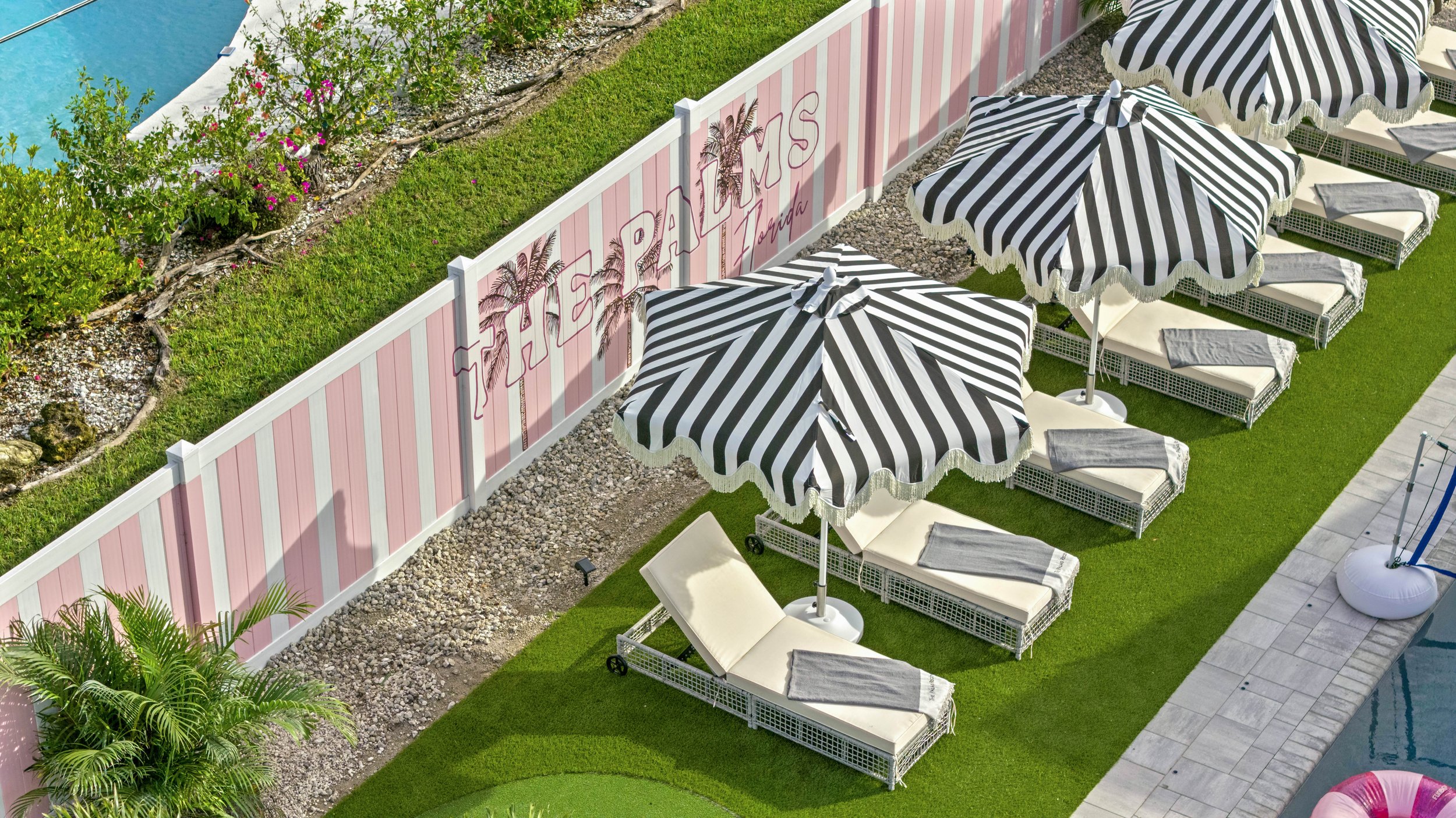 Poolside area with striped black and white umbrellas, white lounge chairs with gray towels, green grass, pink and white striped fence with palm tree illustrations and the words "The Palms Florida," colorful plants, and part of a swimming pool.