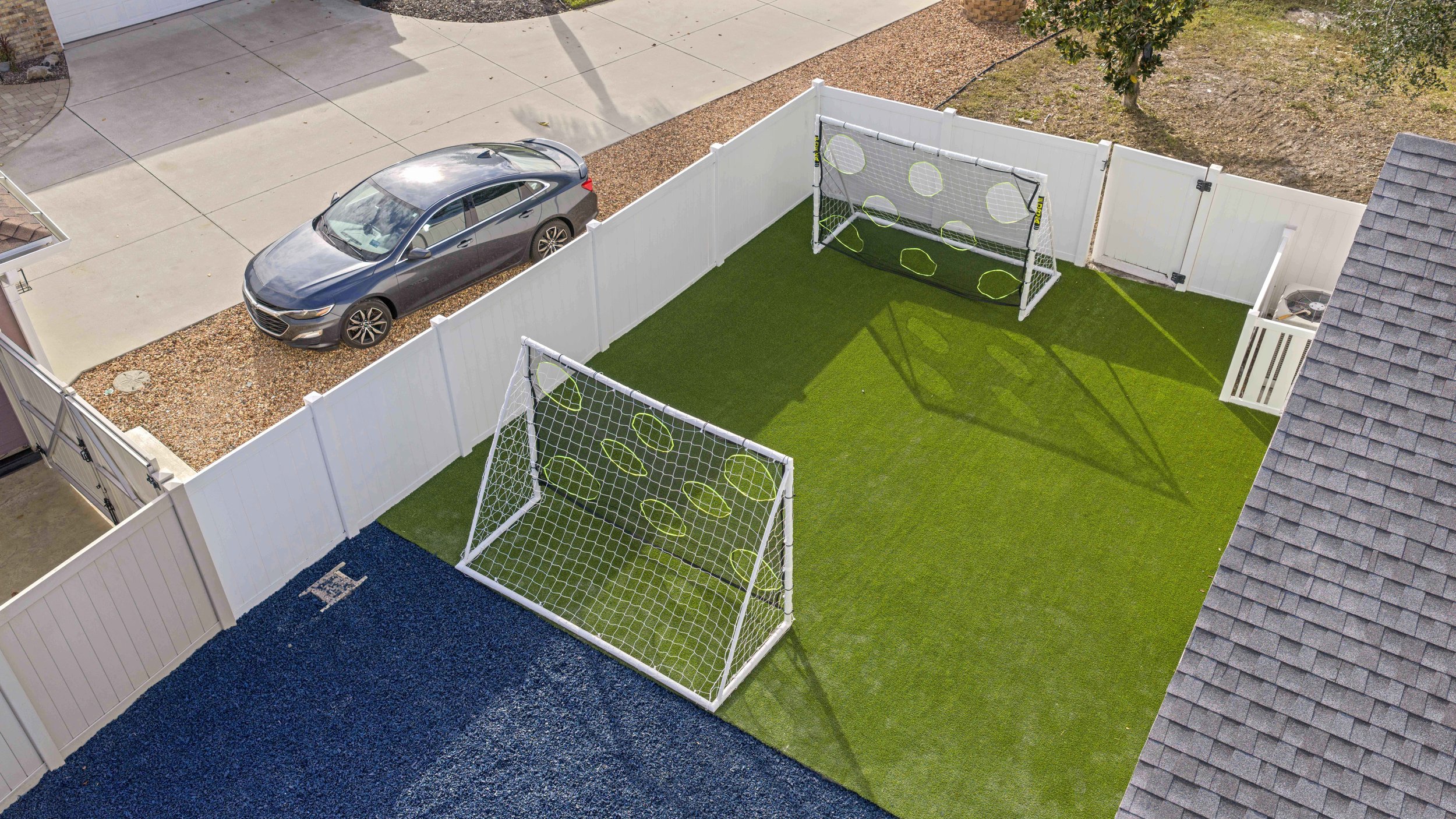 A backyard with artificial grass and two small soccer goals. A white fence surrounds the yard, and a gray car is parked in the driveway outside.