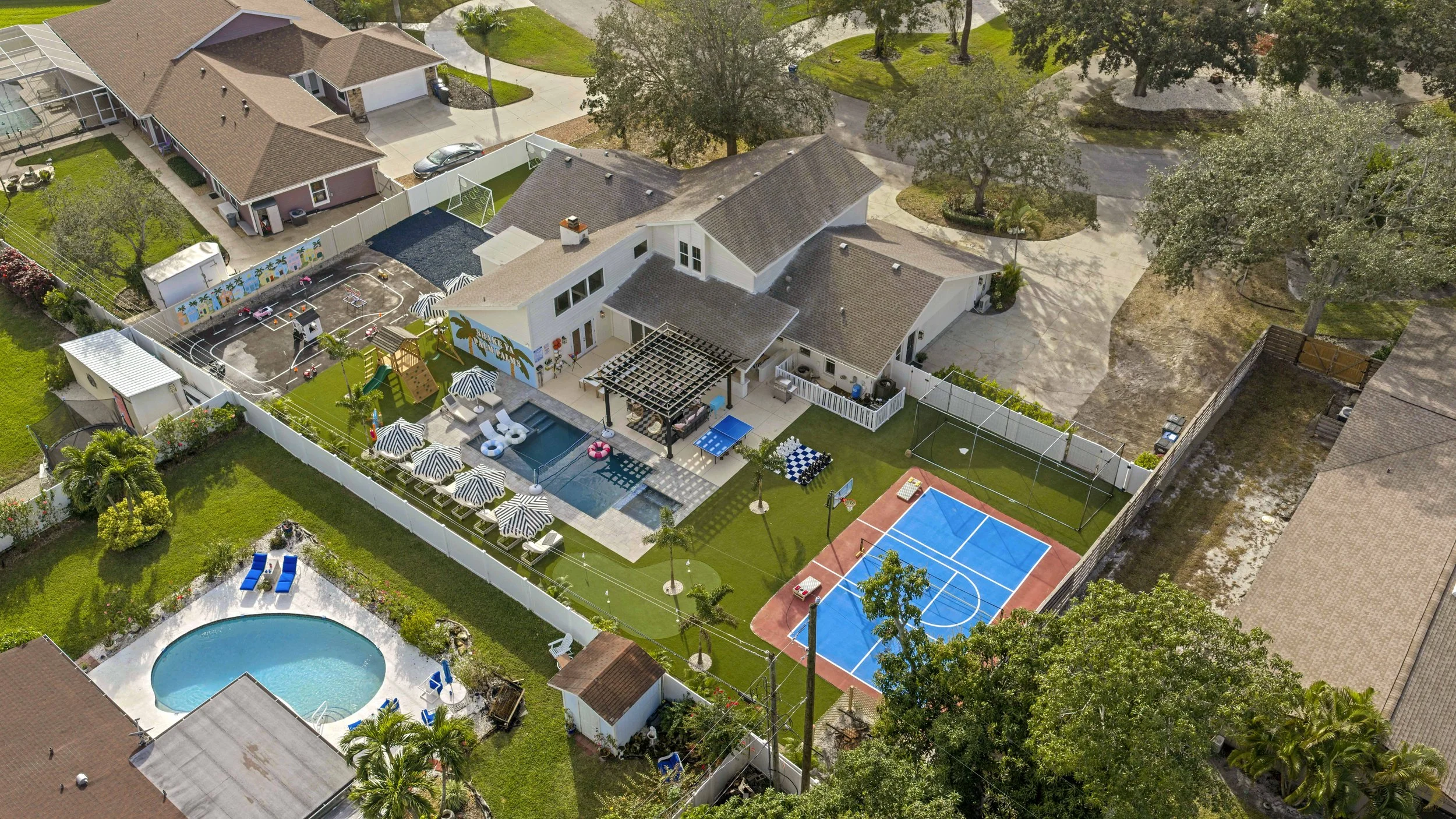 Aerial view of a backyard with a swimming pool, outdoor seating area with umbrellas, a basketball court, and a trampoline, surrounded by a white privacy fence and neighboring houses.