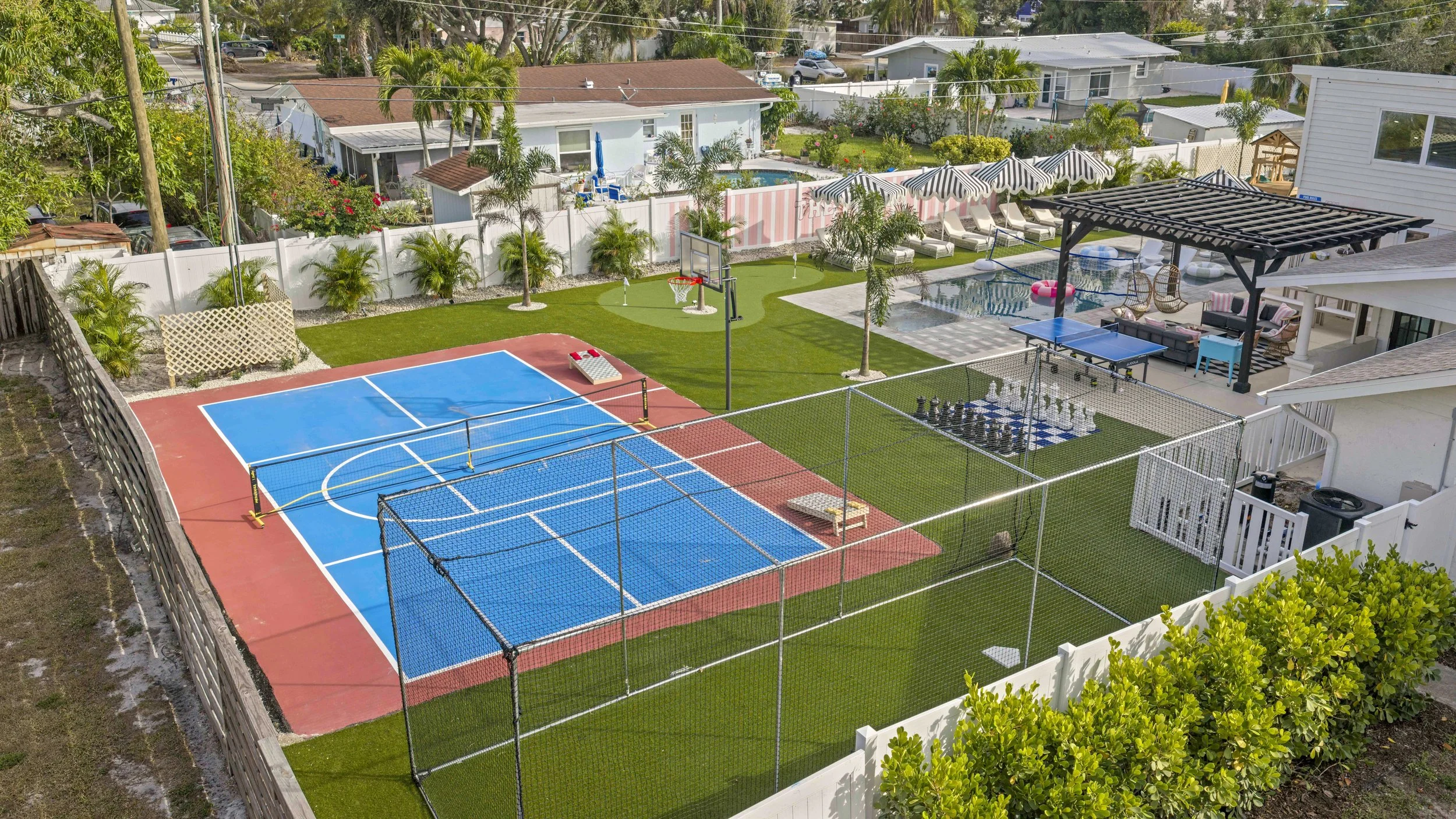 A backyard with a tennis court, a basketball hoop, a small pool, a shaded outdoor seating area, and a chessboard with large chess pieces, surrounded by green grass, palm trees, and a white fence.