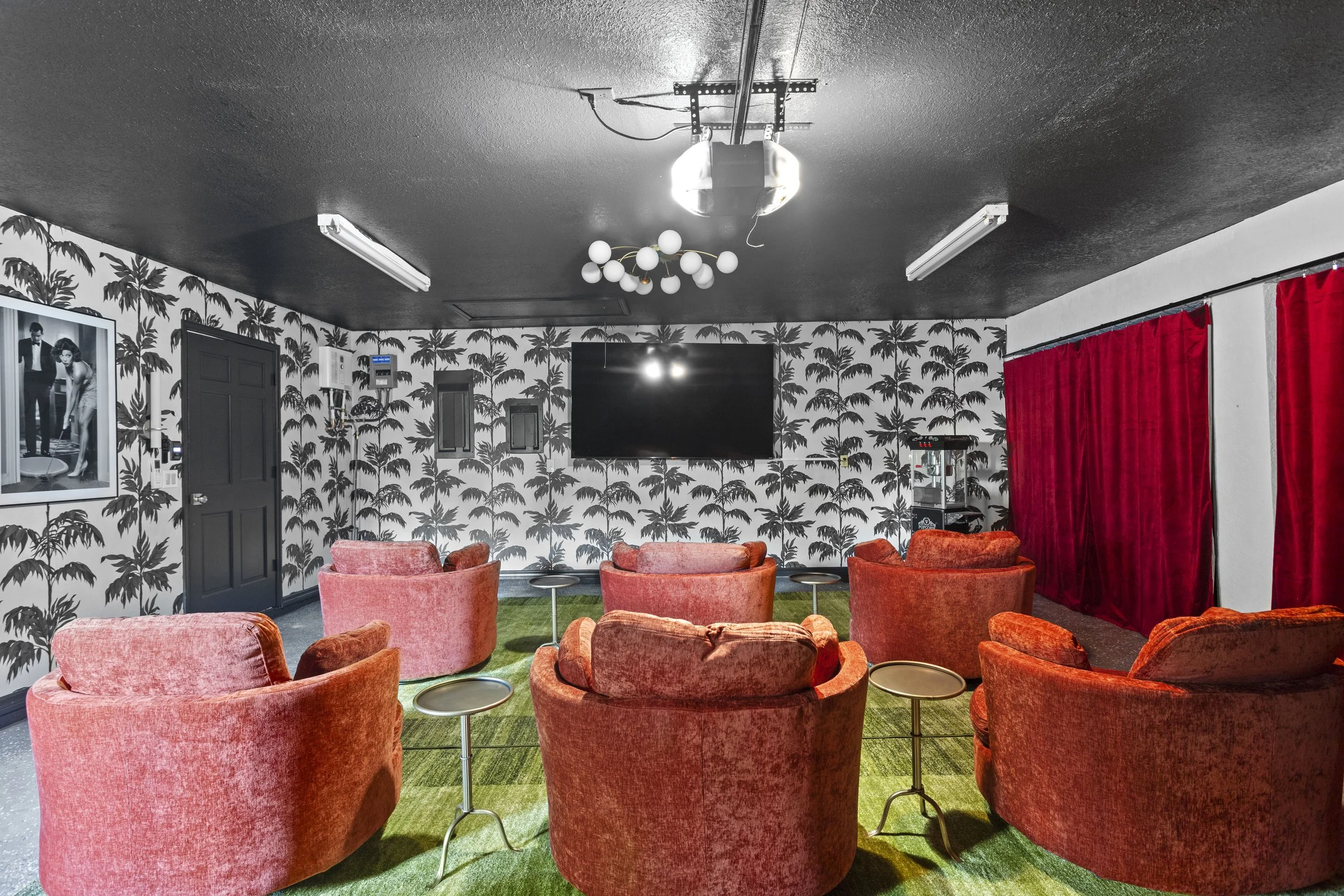 A lounge room with coral velvet armchairs and small round tables on a green carpet, black and white palm tree wallpapered walls, red curtains covering a window, a large television on the wall, and ceiling lights.