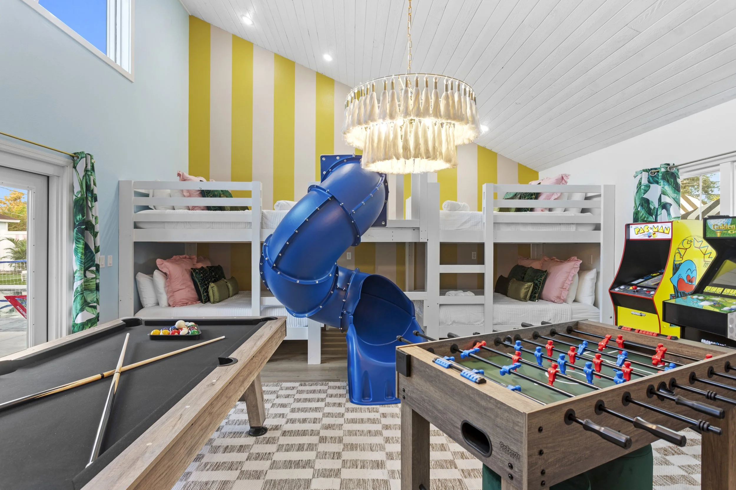 Indoor playroom with two-story bunk bed, blue spiral slide, puzzle table, arcade games, pool table, and large windows with tropical curtains.