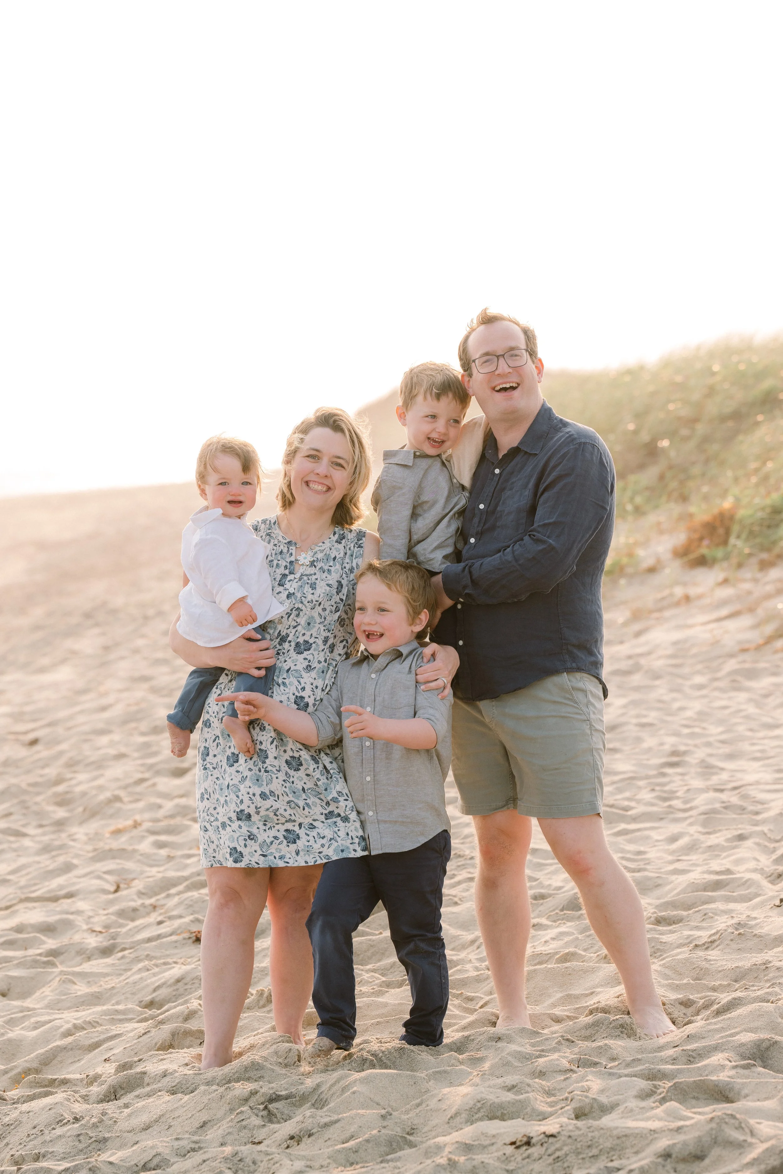 Family of six enjoying a day at the beach, standing on sand with a grassy dune in the background, smiling and laughing together.