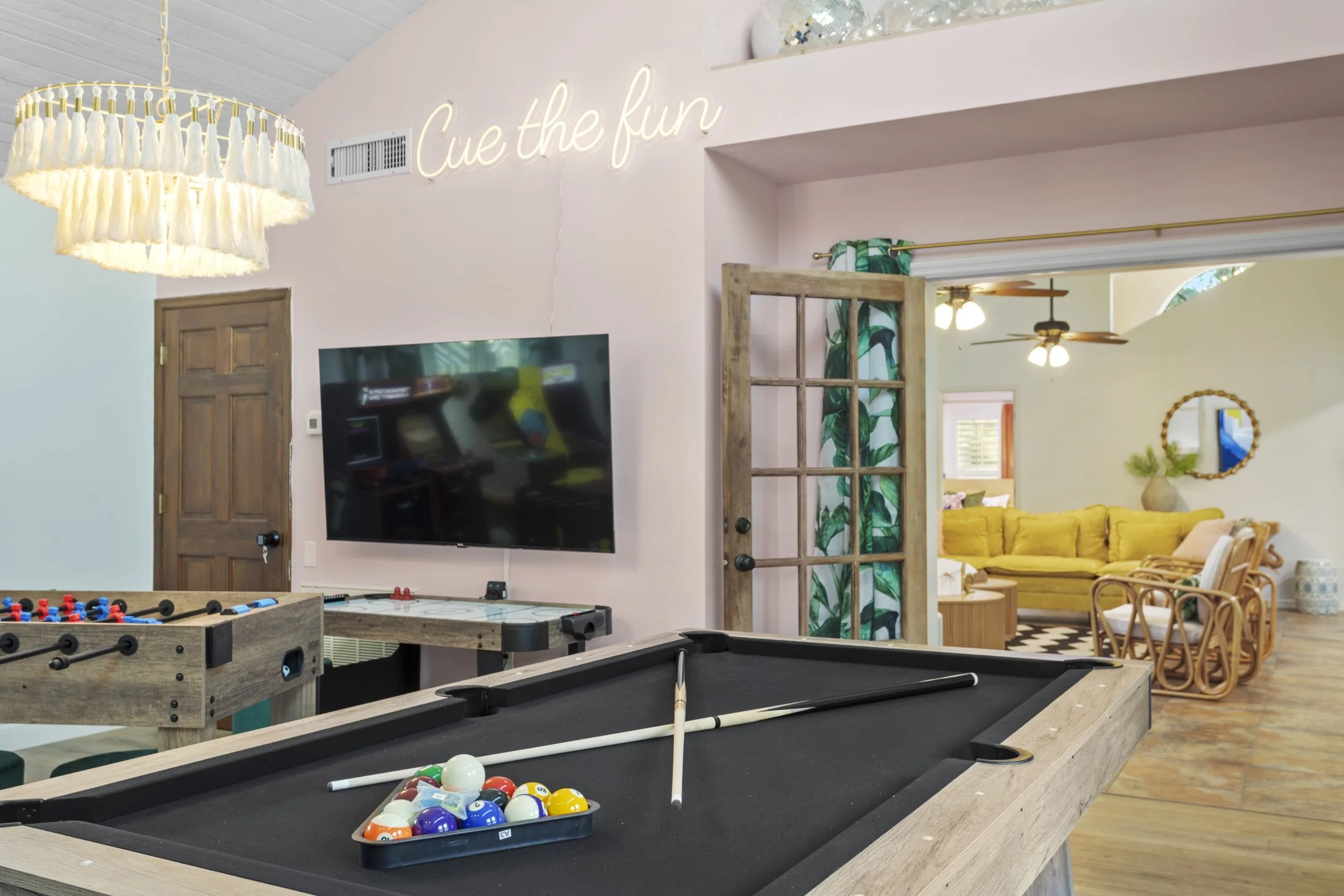 Indoor recreational room with pool table, foosball table, and flat-screen TV on pink wall with neon sign reading "Cue the fun." Open doorway leads to a living room with yellow sofa and rattan chairs.