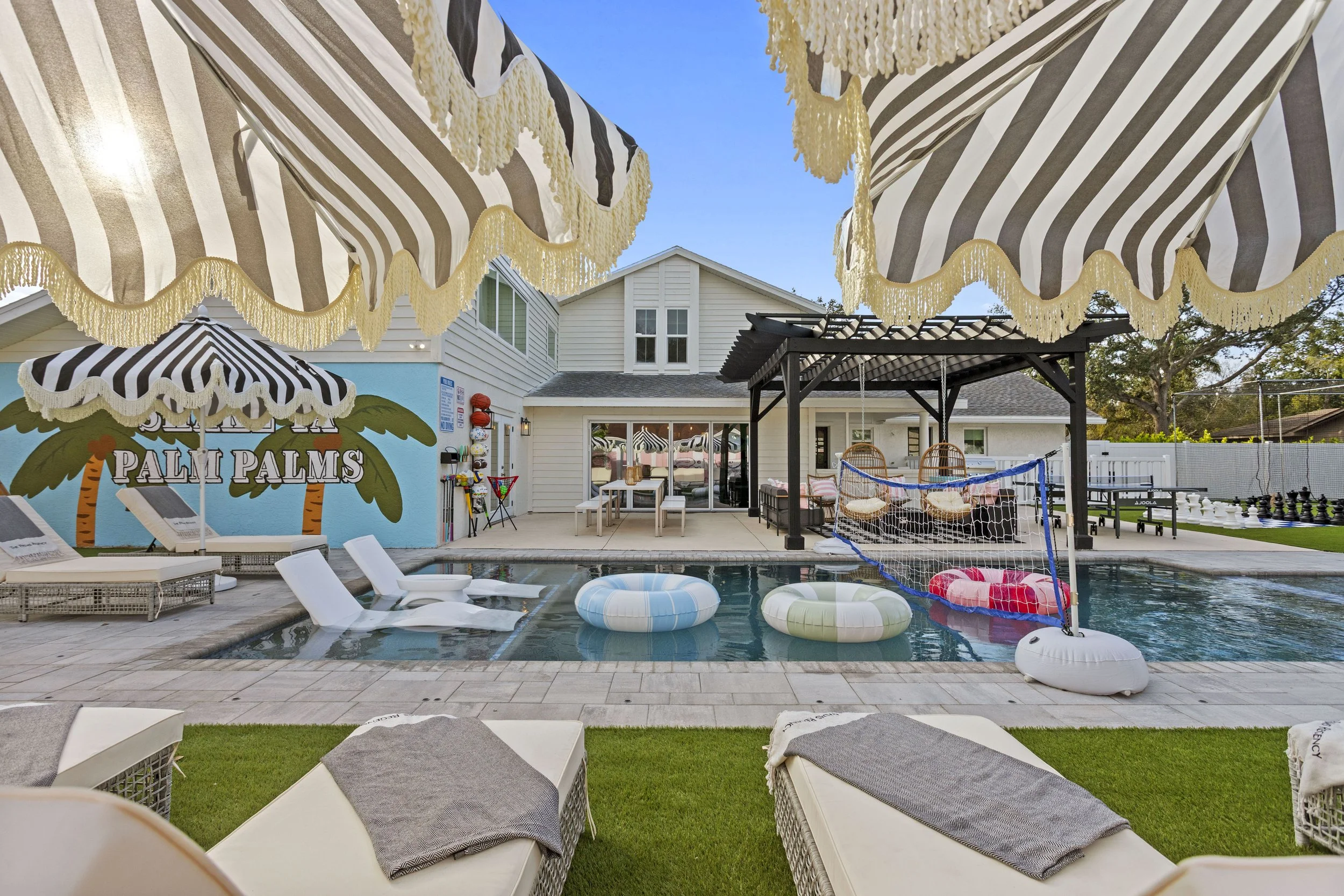 Backyard with a pool, lounge chairs with towels, umbrellas with fringes, a shaded pavilion, colorful floating pool rings, a volleyball net, and a chess set, with a white house and blue sky in the background.
