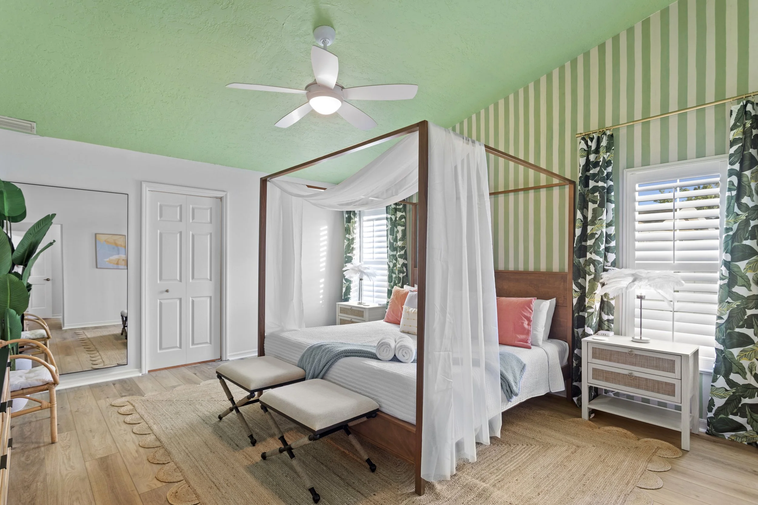 Bedroom with a four-poster bed, white canopy, white and orange pillows, blue throw blanket, and rolled towels. The room has green striped and tropical-patterned curtains, a white nightstand with a lamp, a ceiling fan, and wood flooring with a woven rug.