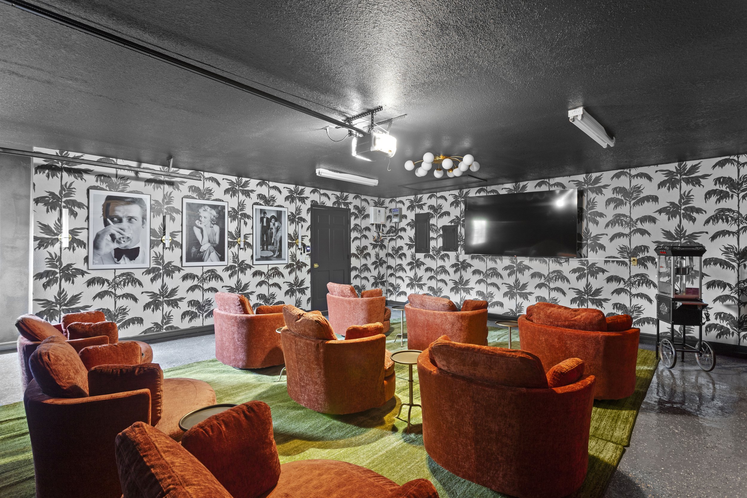 A retro-style lounge room with orange velvet armchairs on a green patterned carpet, black and white palm tree wallpaper, framed black and white photos on the wall, a large flat-screen TV, and various light fixtures on the ceiling.