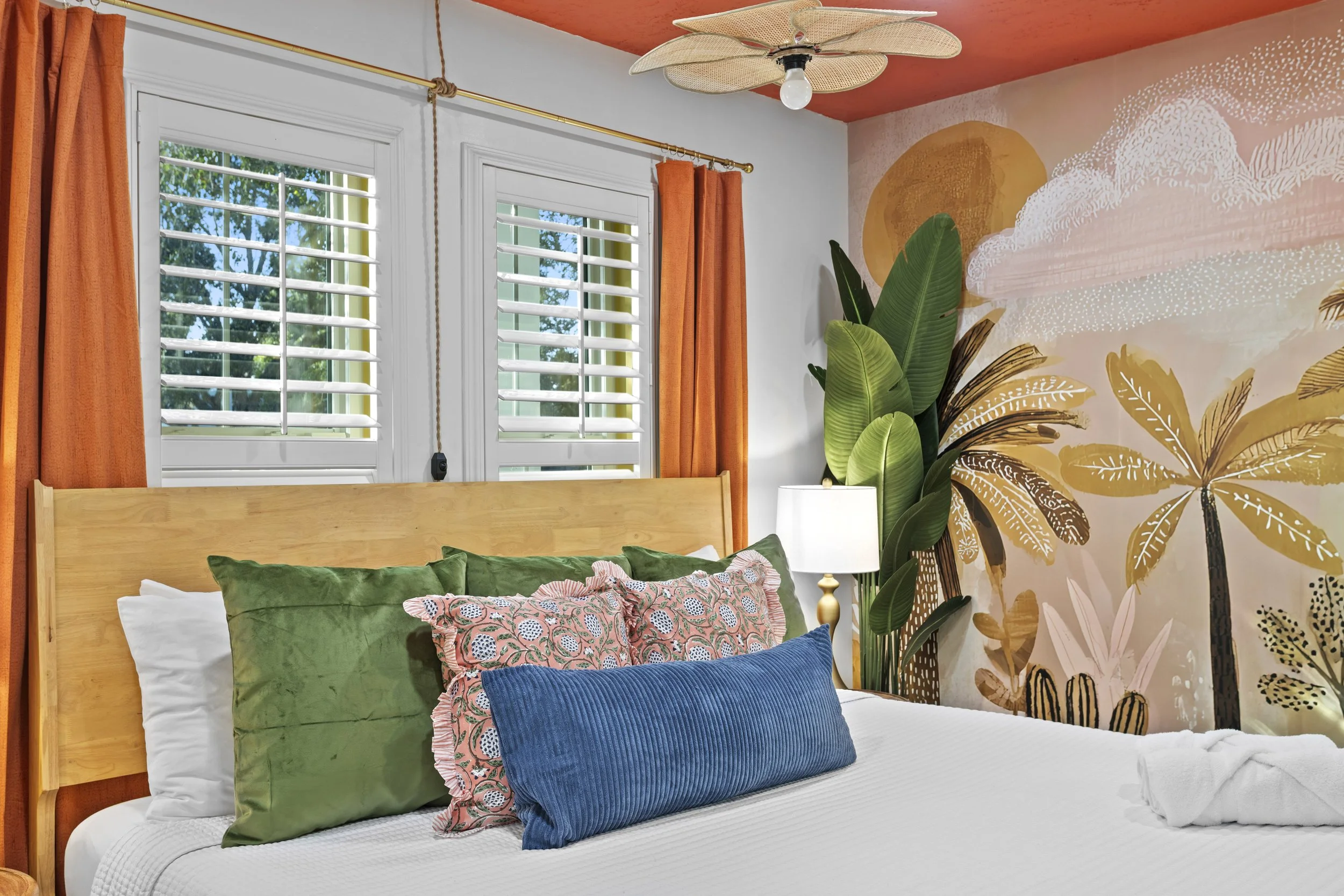 A bedroom with a wooden bed frame, white bedding, green, pink, and blue throw pillows, orange curtains, a wall mural of trees and clouds, a large potted plant, and a bedside lamp.