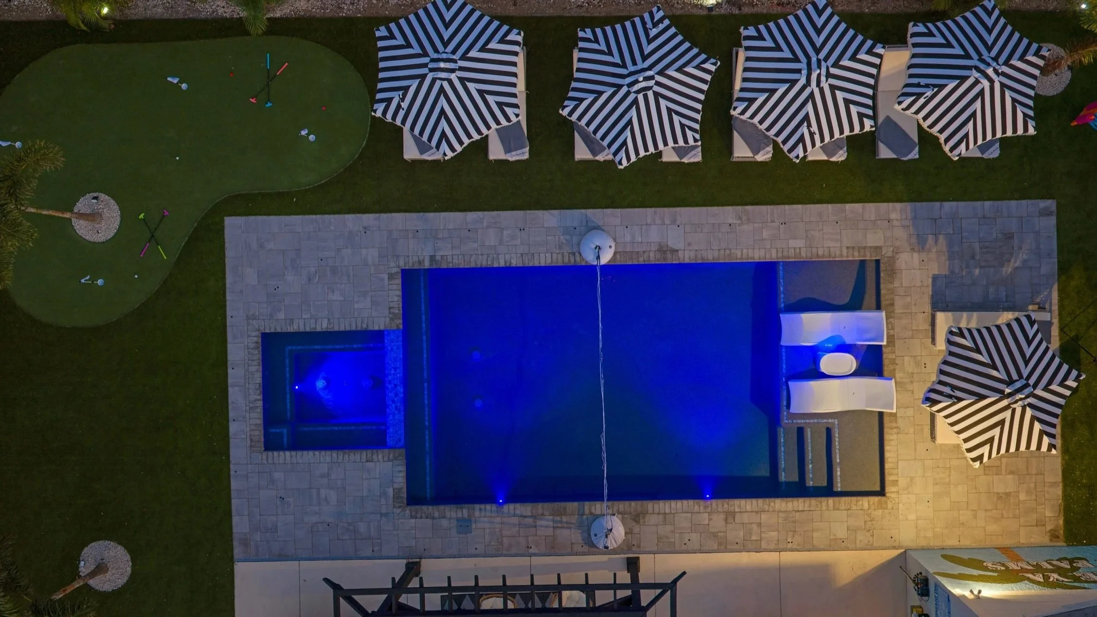 Aerial view of a backyard pool illuminated with blue lights, surrounded by a paved deck area with seating, striped umbrellas, and a grassy area with a putting green and decorative golf club markers.