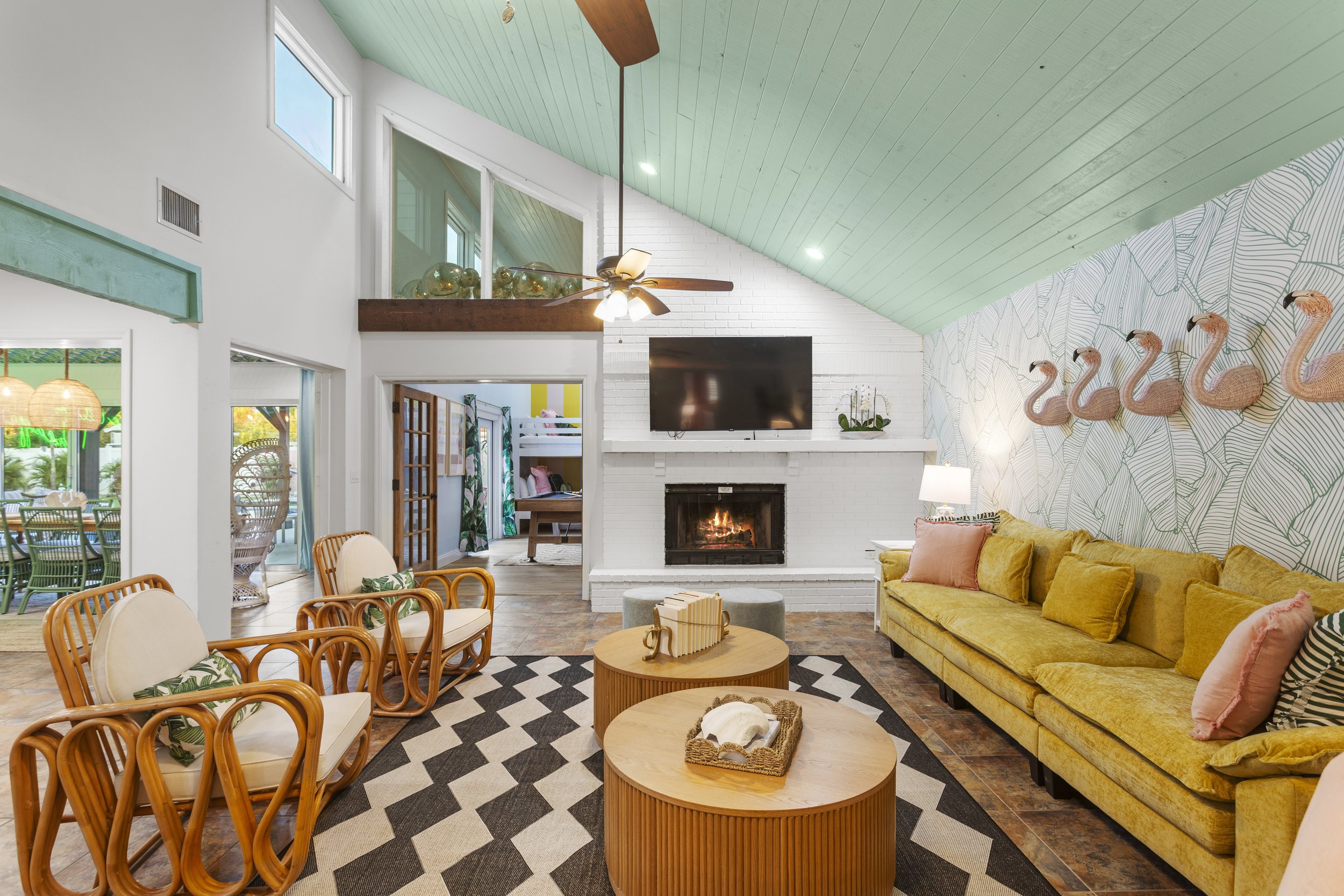 Living room with white brick fireplace, yellow sofa, wall art of pink flamingos, wooden coffee tables, patterned rug, green ceiling, and large windows.