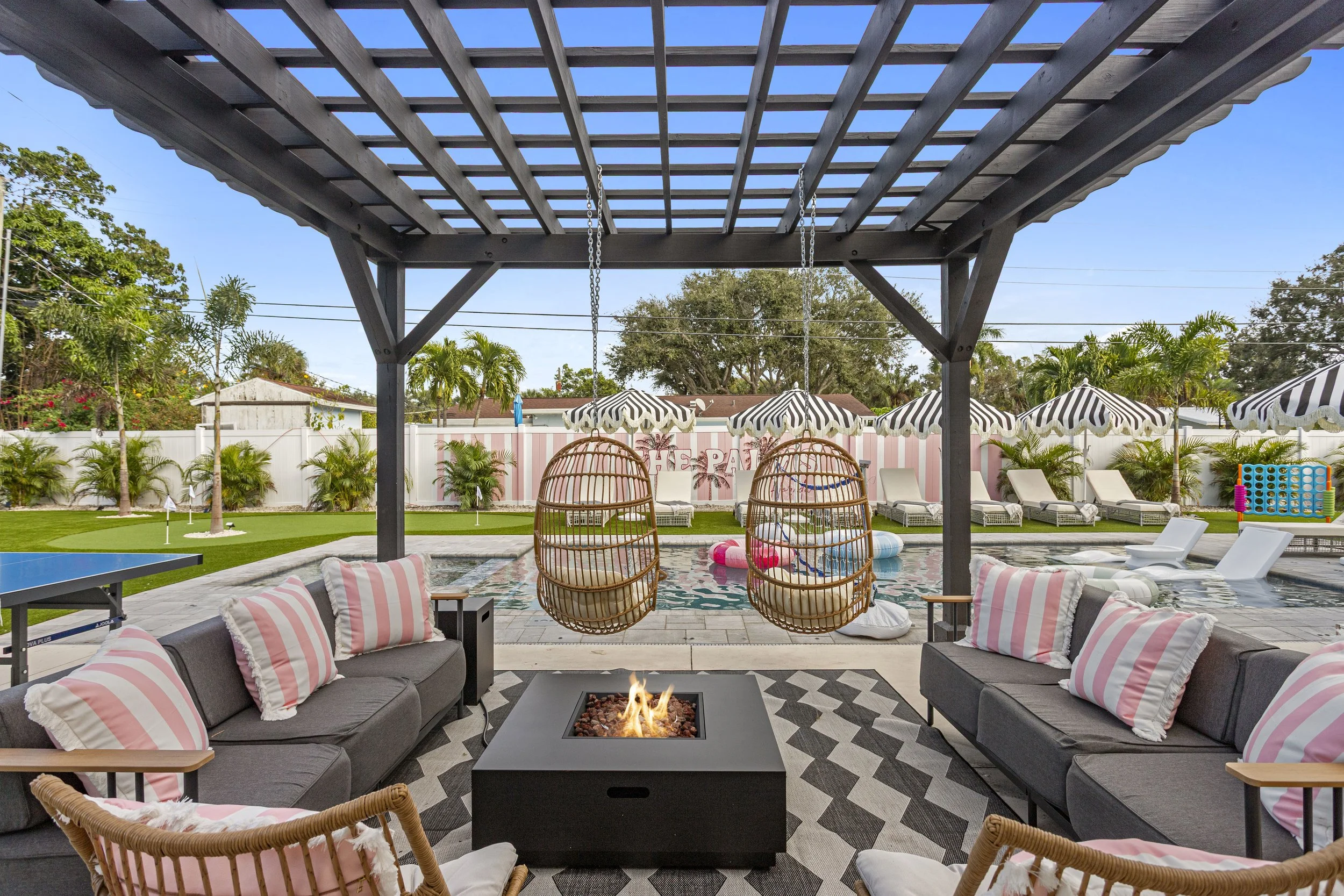 Outdoor patio with seating area, hanging wicker chairs, and pool with lounge chairs and umbrellas in the background.