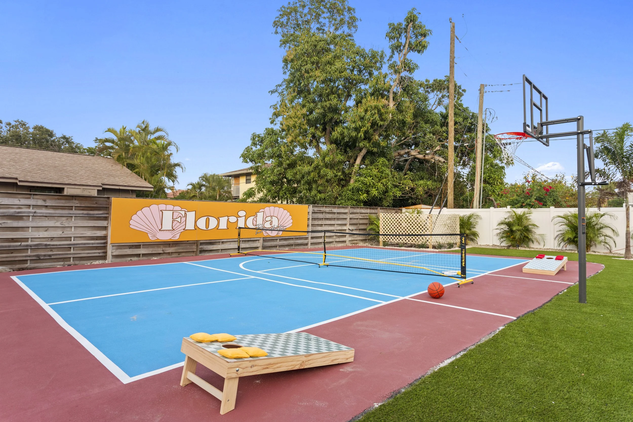 A backyard basketball court with a mini volleyball net in the foreground, a basketball resting on the court, a cornhole game to the right, and a large sign that says 'Florida' with seashell graphics on a wooden fence. There are trees and a blue sky in the background.