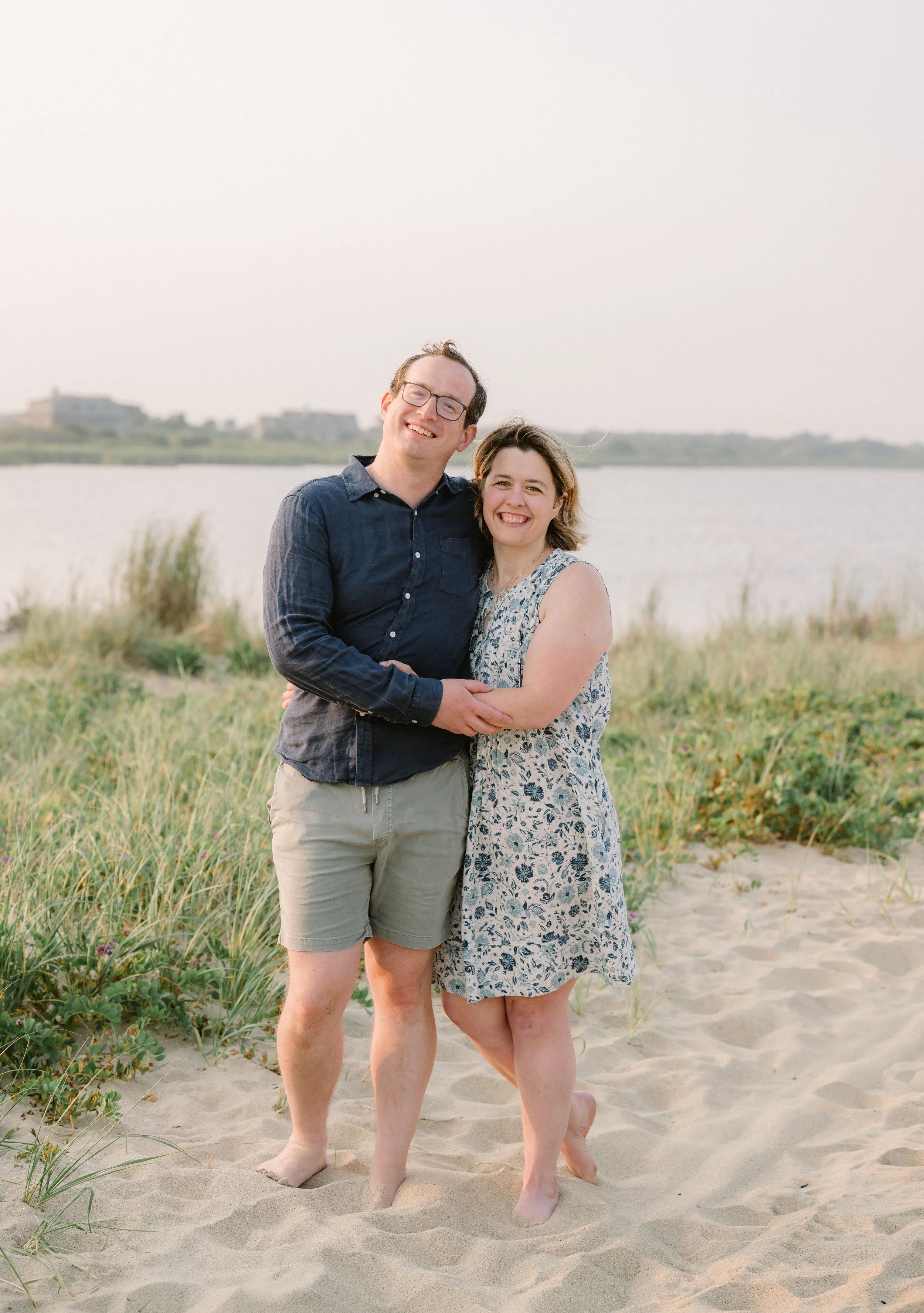 A smiling man and woman standing close together on a sandy beach with green grass, near a body of water, with houses in the background during what appears to be sunset.